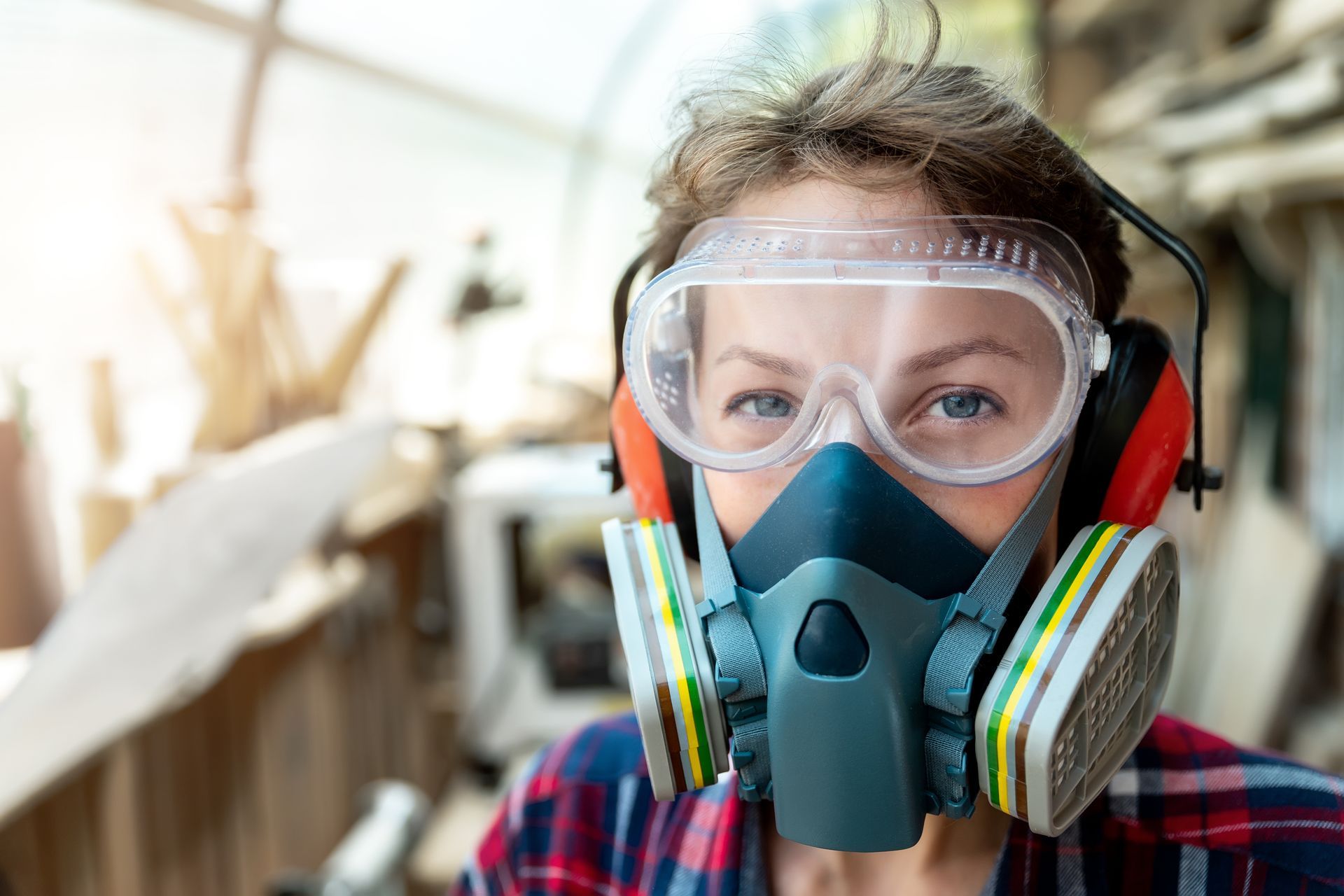 Person wearing safety goggles, respirator mask, and earmuffs in a workshop with woodworking tools.