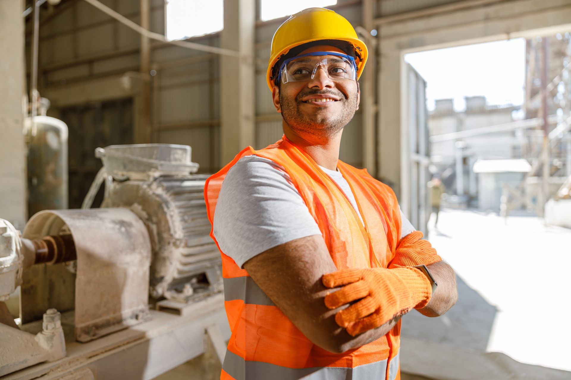 Factory worker in hard hat and orange vest standing beside industrial machinery, smiling with crossed arms.