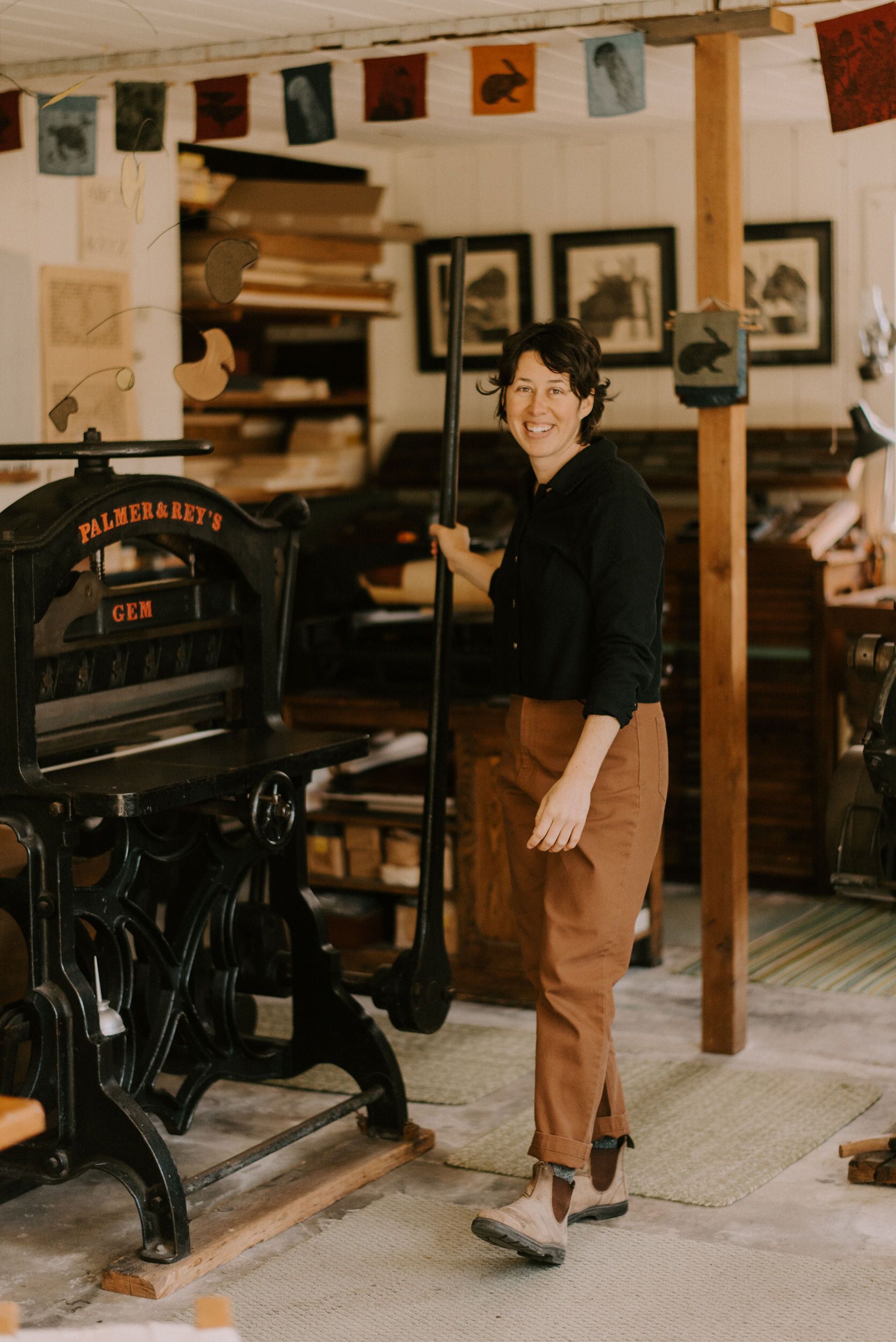 Person standing in a vintage print shop beside an old black press, smiling at the camera.