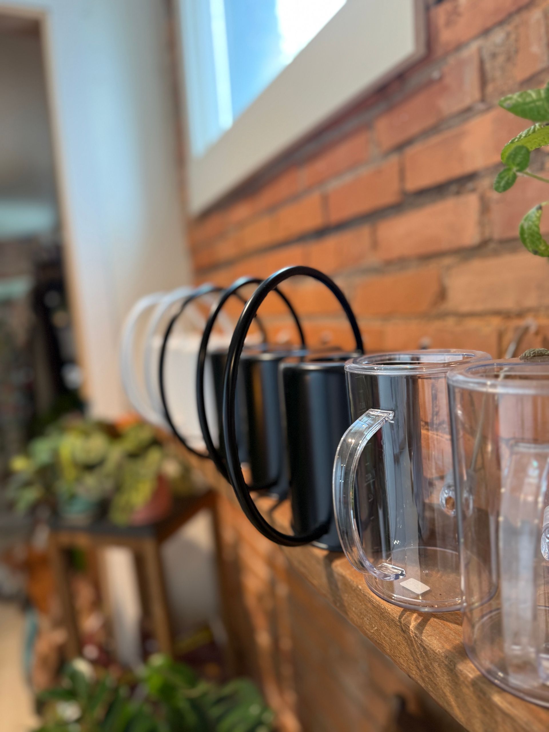 Clear mugs hanging on black wall hooks beside a brick wall and window.
