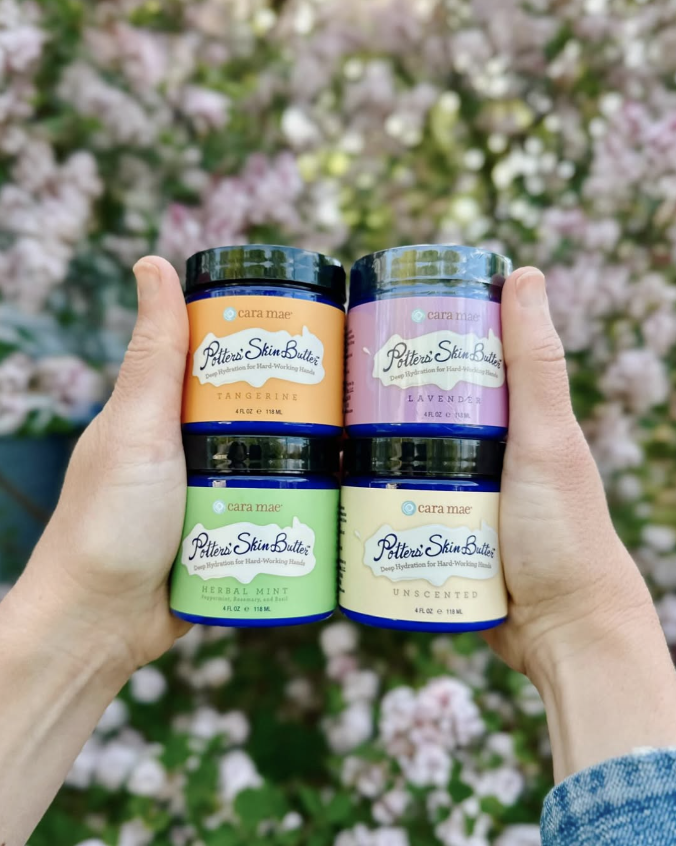 Hands holding four small scented candle jars with pastel labels against a blurred floral background