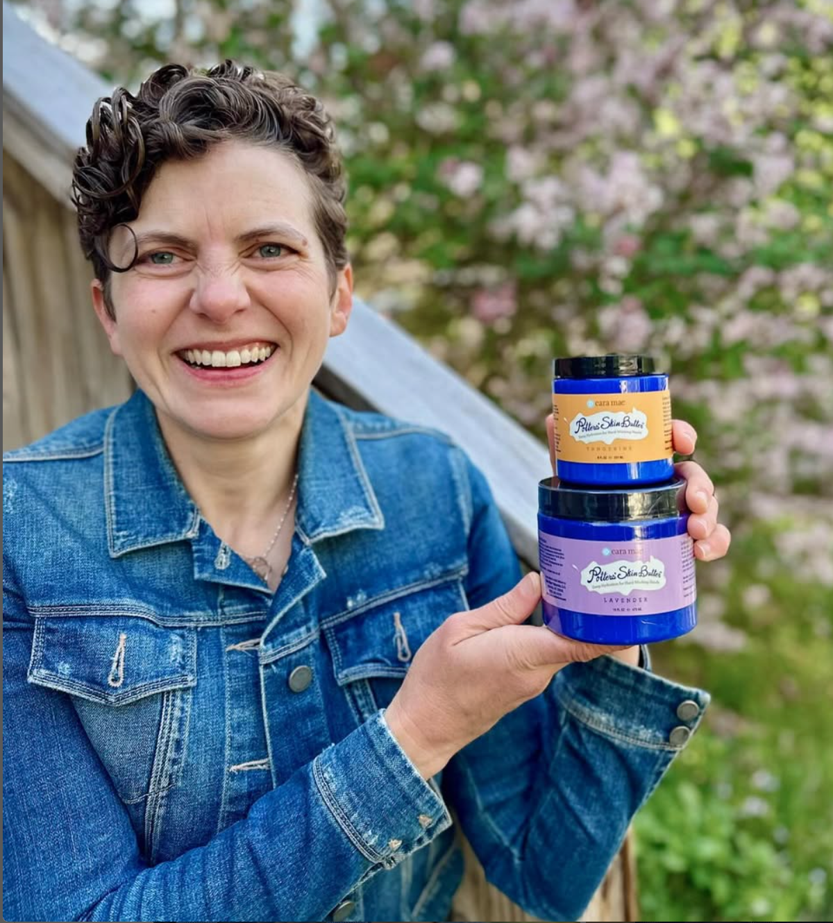 Smiling person in a denim jacket holding two blue-labeled jars outdoors