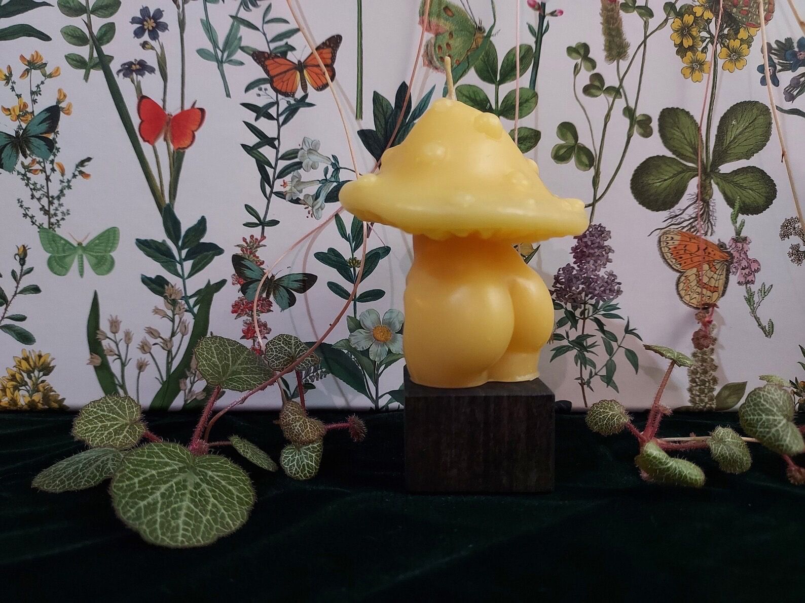 Yellow mushroom-shaped candle on a dark base before butterfly-patterned wallpaper and leaves