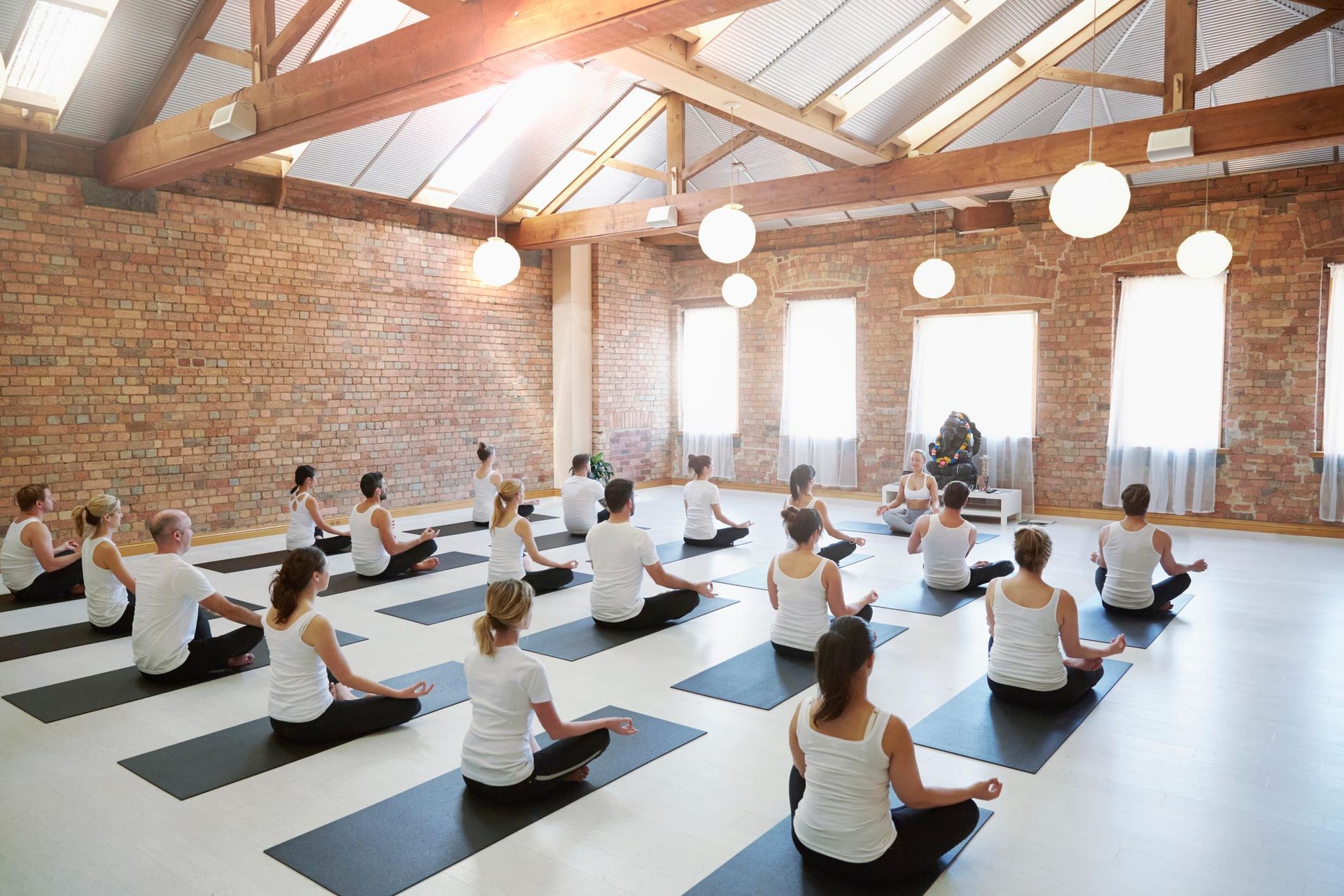 People in a yoga class, meditating on mats in a bright studio with exposed brick and skylights.