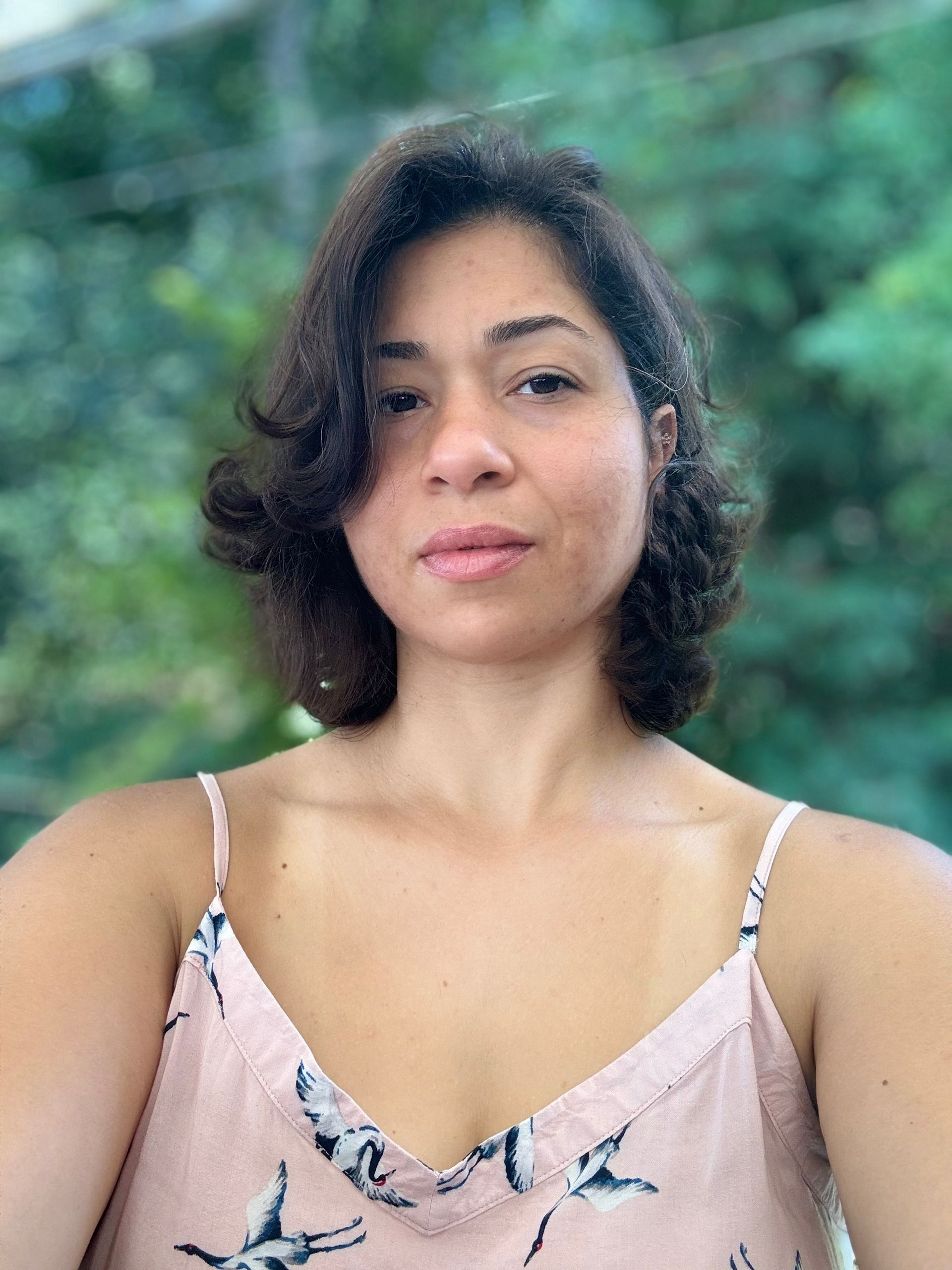 Woman with dark, wavy hair, wearing a pink top with a bird pattern, looking at the camera. Outdoors with trees.