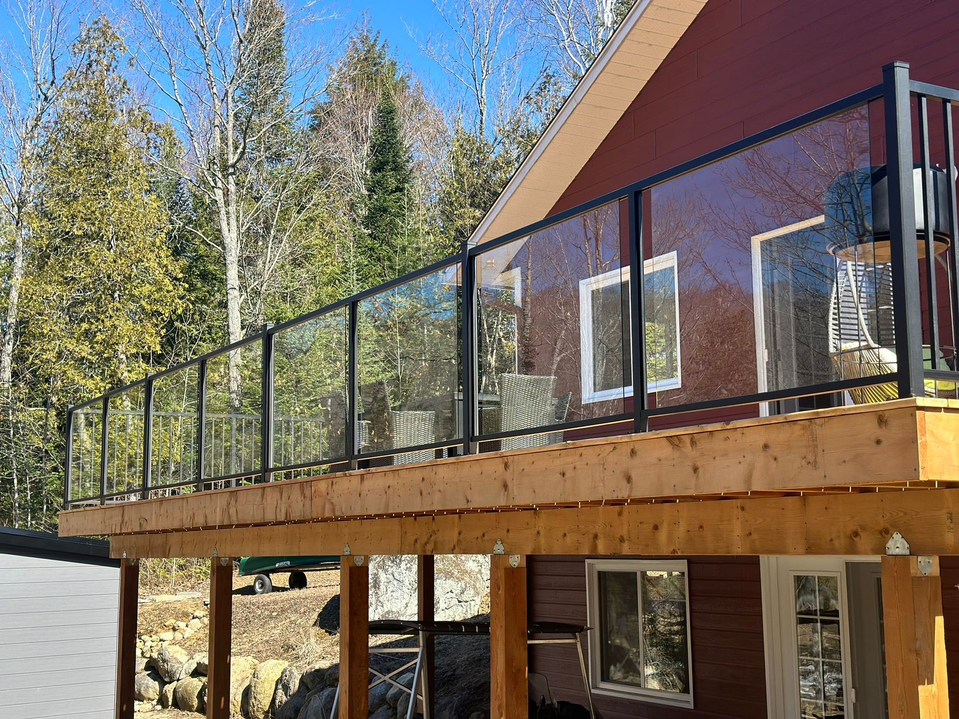 Une terrasse en bois avec une balustrade en verre et une maison rouge en arrière-plan.
