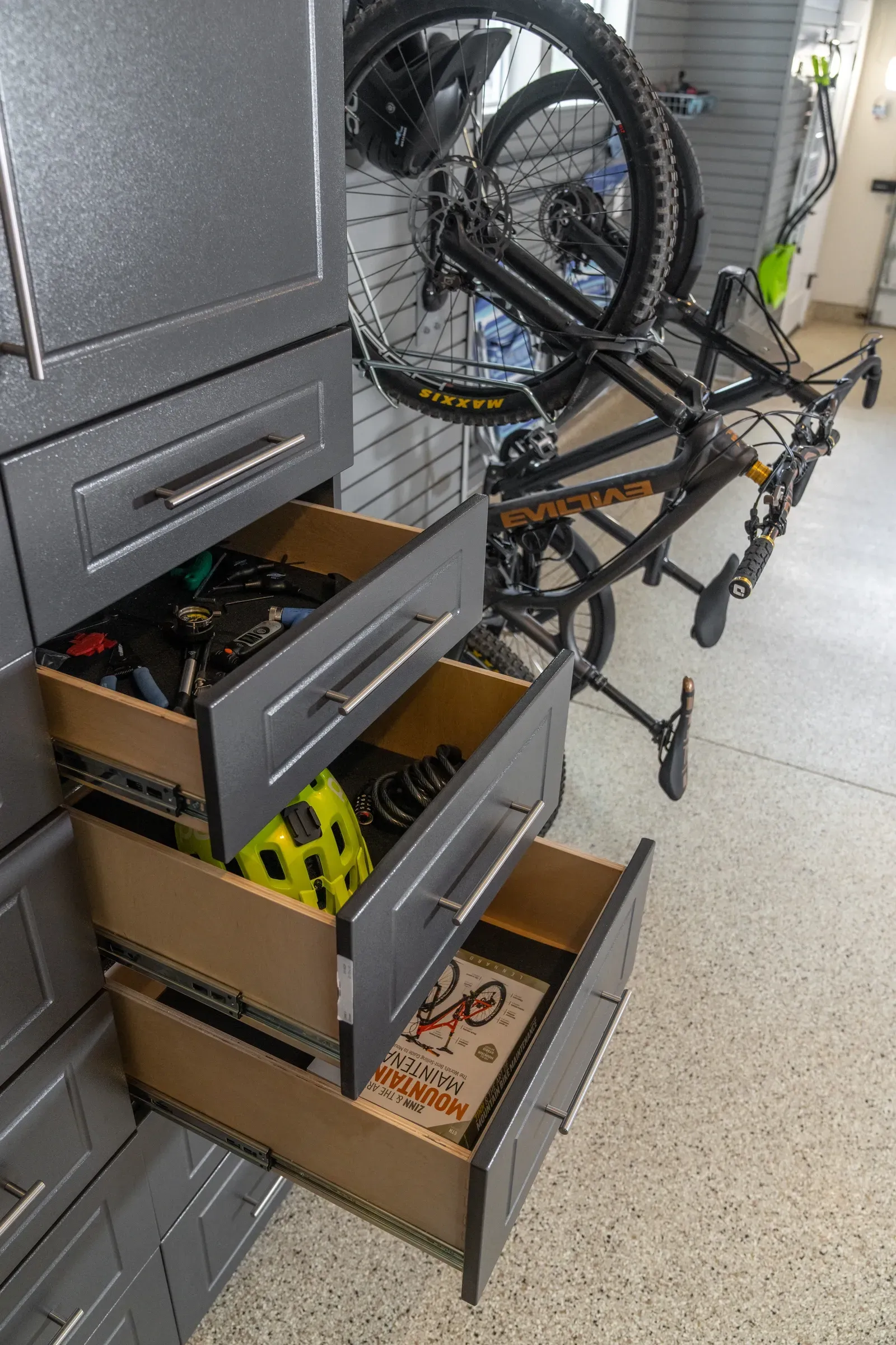 Open drawers in a gray cabinet with bike parts and a bicycle leaning beside it in a garage.
