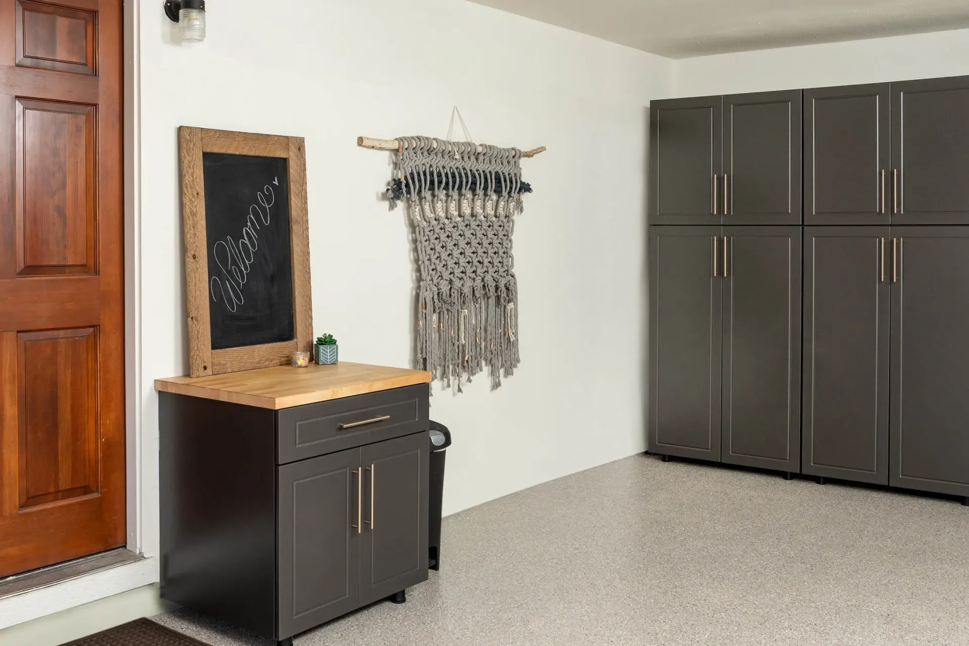 Interior mudroom with black cabinets, small bench, wall mirror, and woven wall hanging on a light wall.