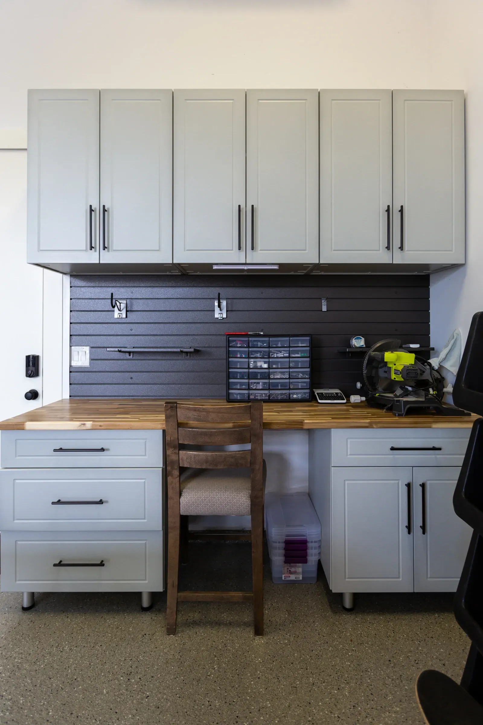 A gray wall cabinet and workbench with drawers, a wooden stool, and tools in a garage workspace.