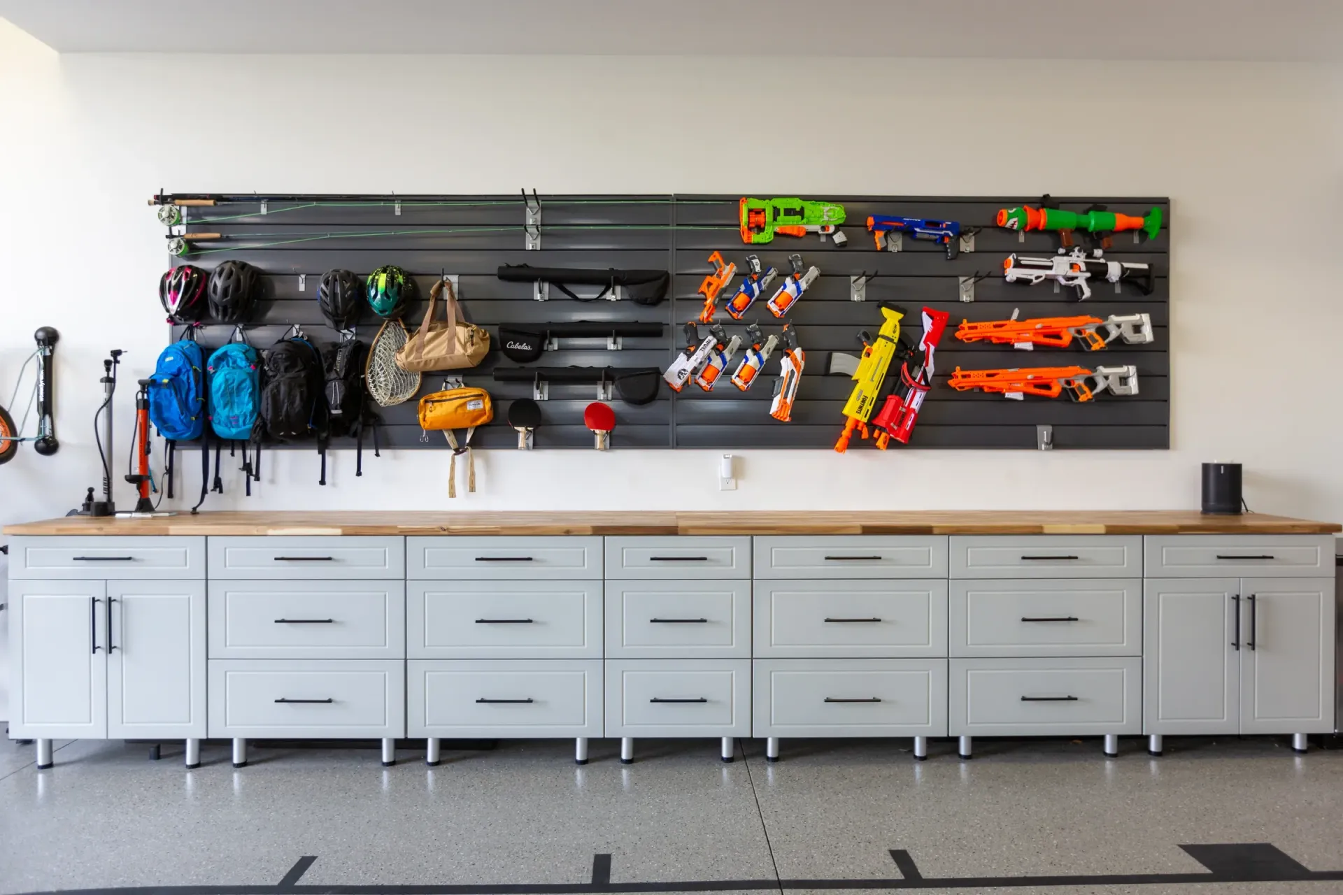 Garage storage wall with organized tools, backpacks, and toy blasters above a long white cabinet.
