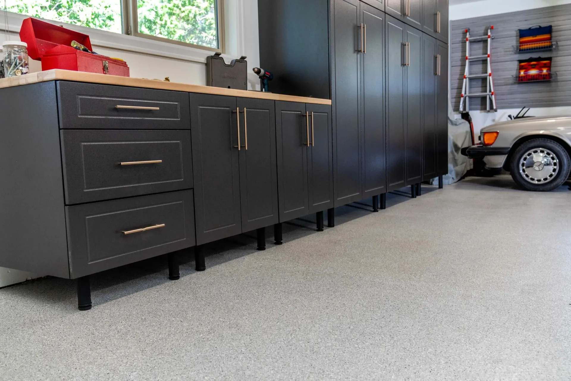 Black garage cabinets and drawers along a speckled floor, with a car and ladder in the background.