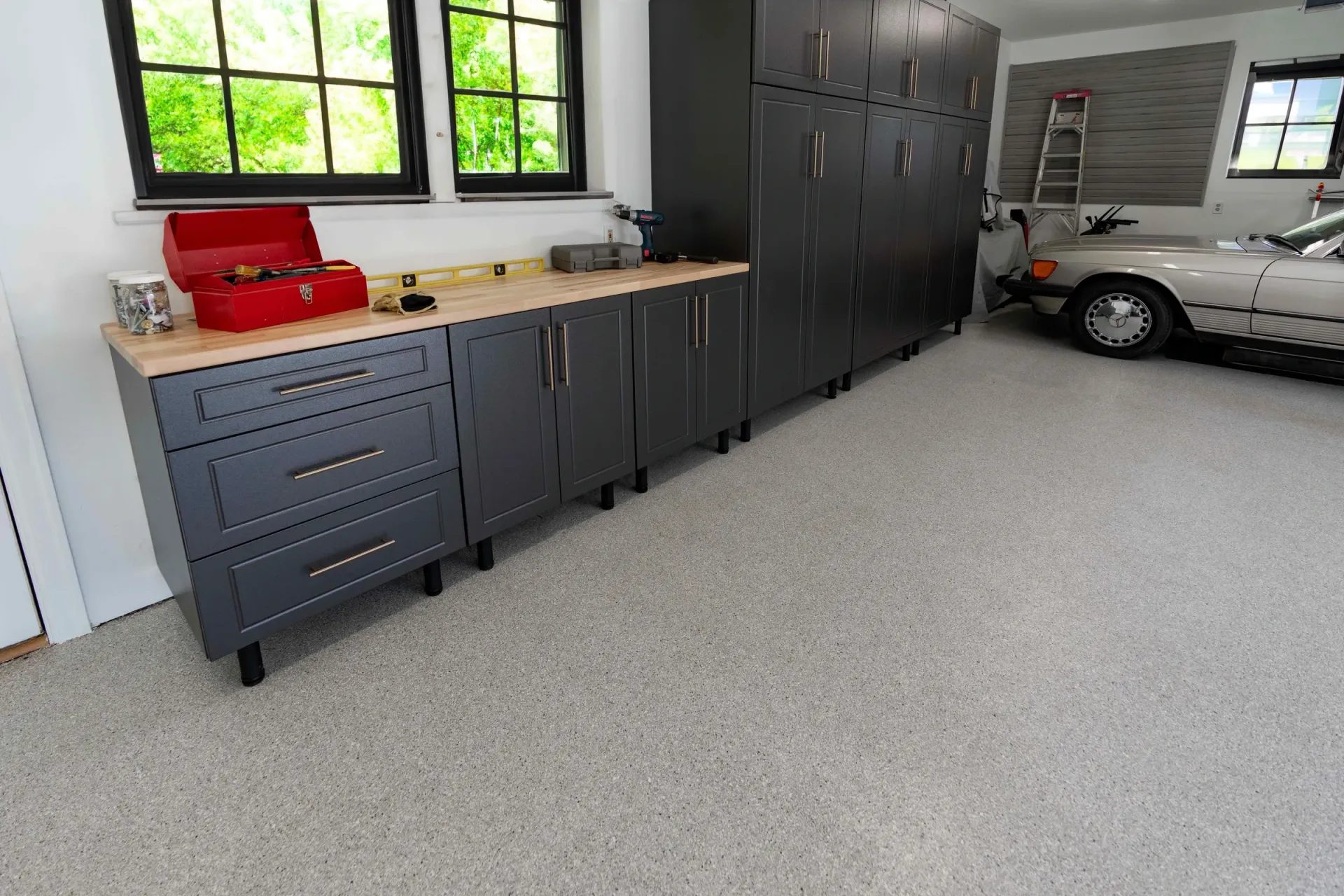 Gray garage cabinets and tool storage along a wall beside a parked car, with a window and speckled floor.