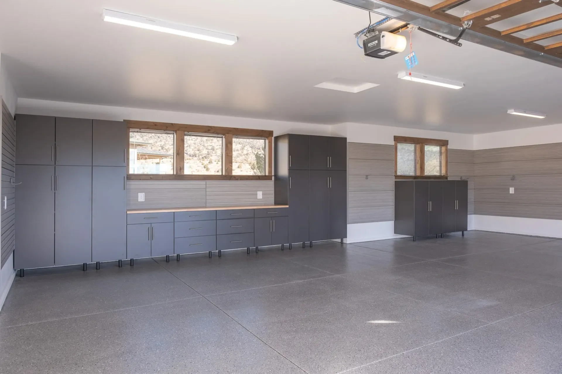 Empty garage with gray cabinets, speckled floor, windows, and overhead lights.