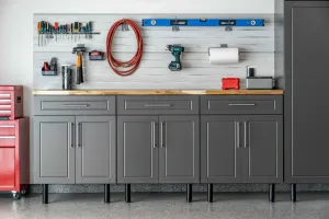 Gray garage workbench with tools, pegboard, red hose, and overhead shelf on a light wall.