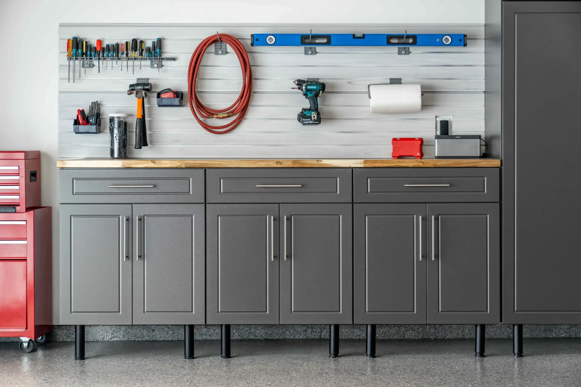 Gray garage workbench with tools, coiled hose, level, and cabinets against a slat wall