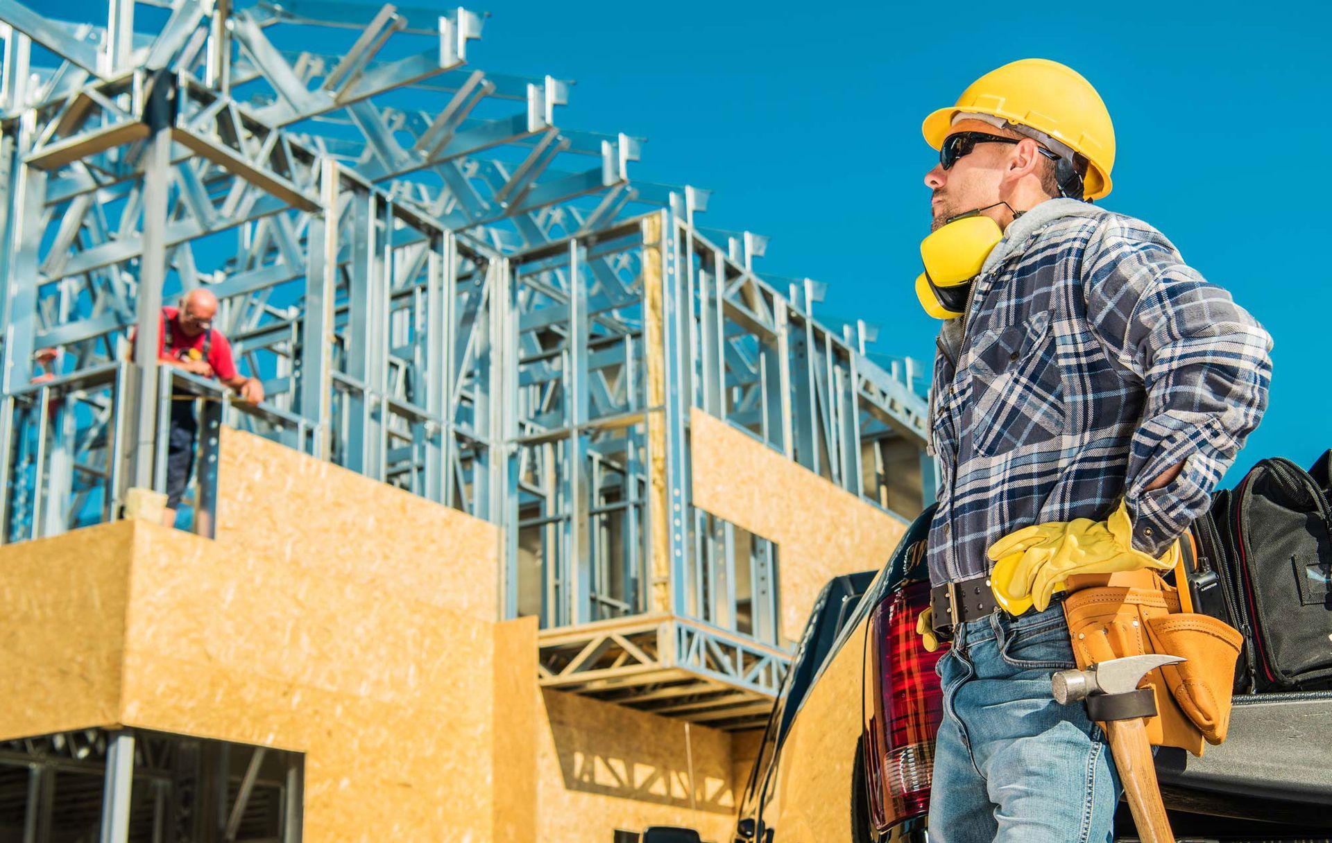Construction worker in yellow hard hat and respirator stands at a wood-framed building site.