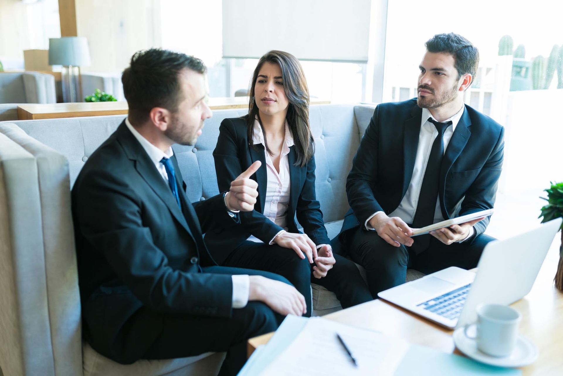 Three business professionals in suits discussing around a laptop in a bright office lounge.