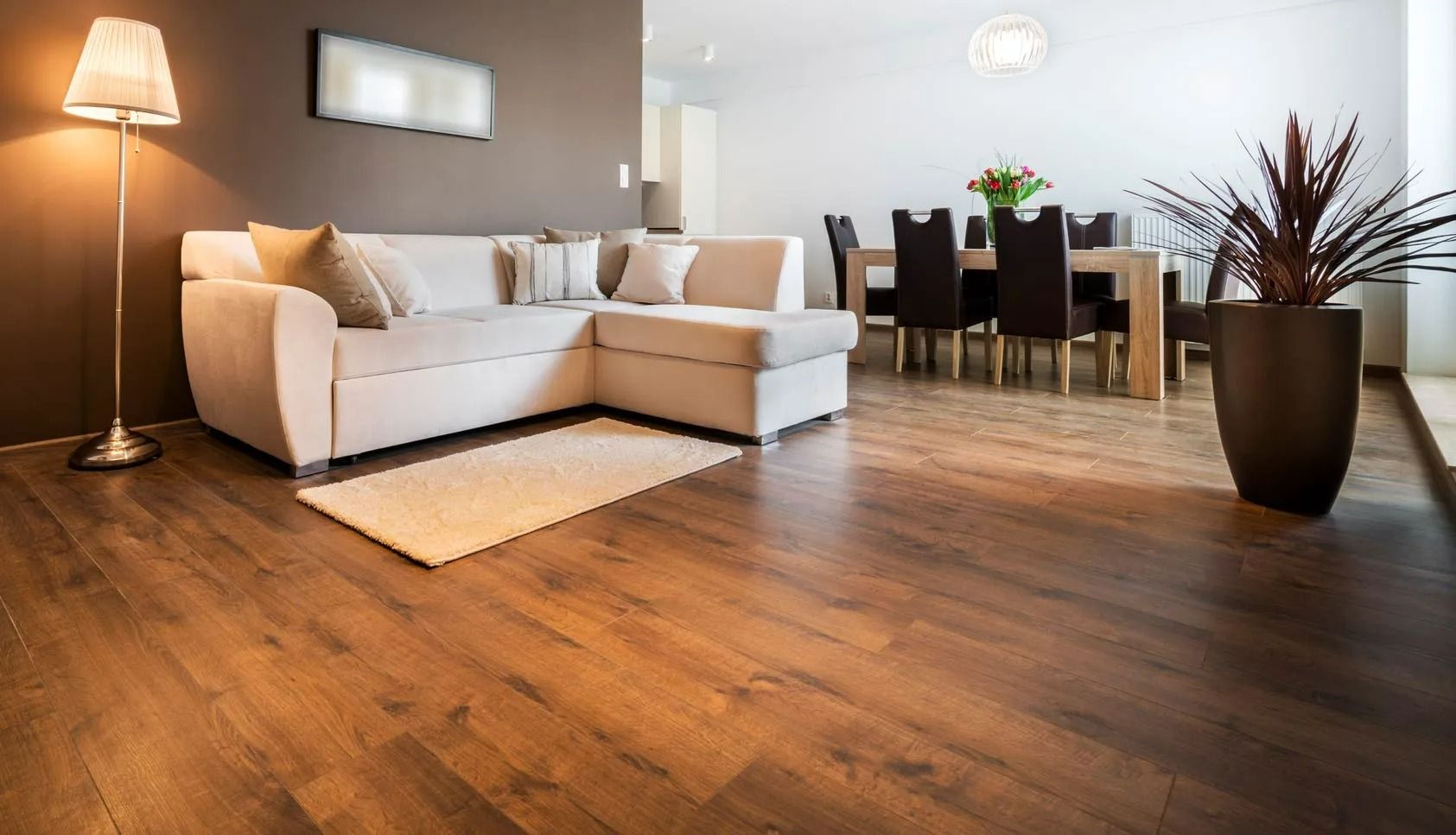 A bright living room features a beige sectional sofa, a floor lamp, and a dining area with dark chairs on wood flooring.