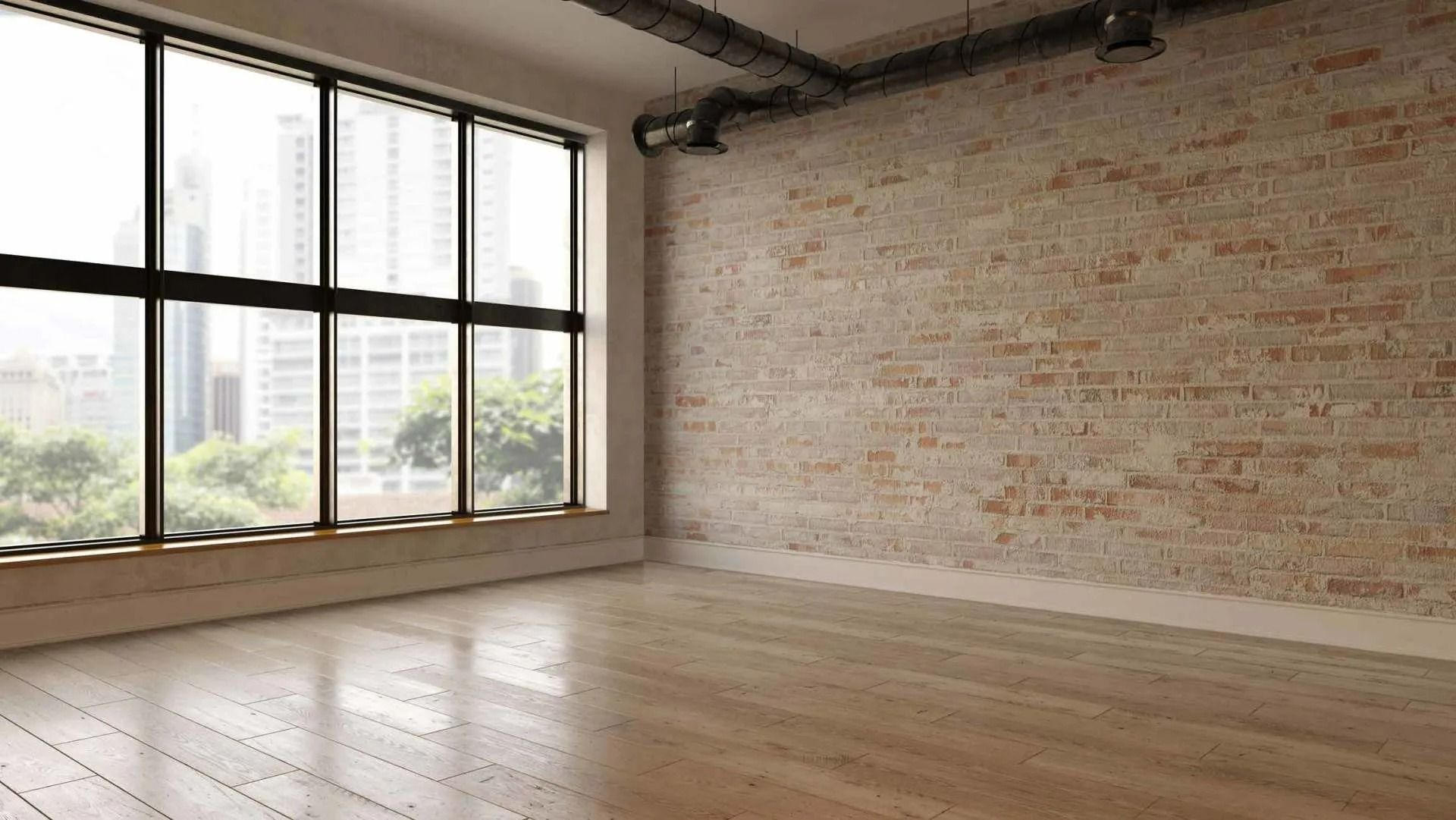 An empty room with a light brick wall, wooden flooring, and large windows overlooking a city skyline.