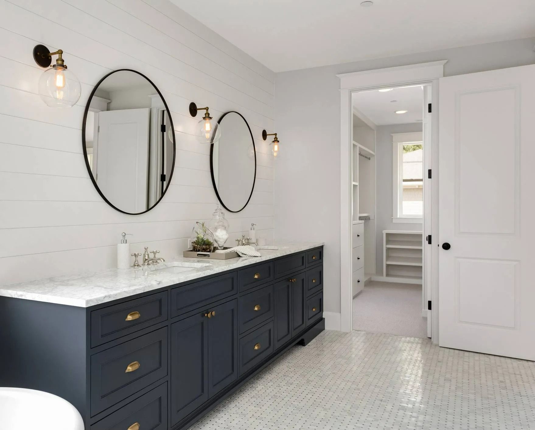 A bright bathroom with a navy double vanity, marble countertop, two oval mirrors, and white shiplap walls.