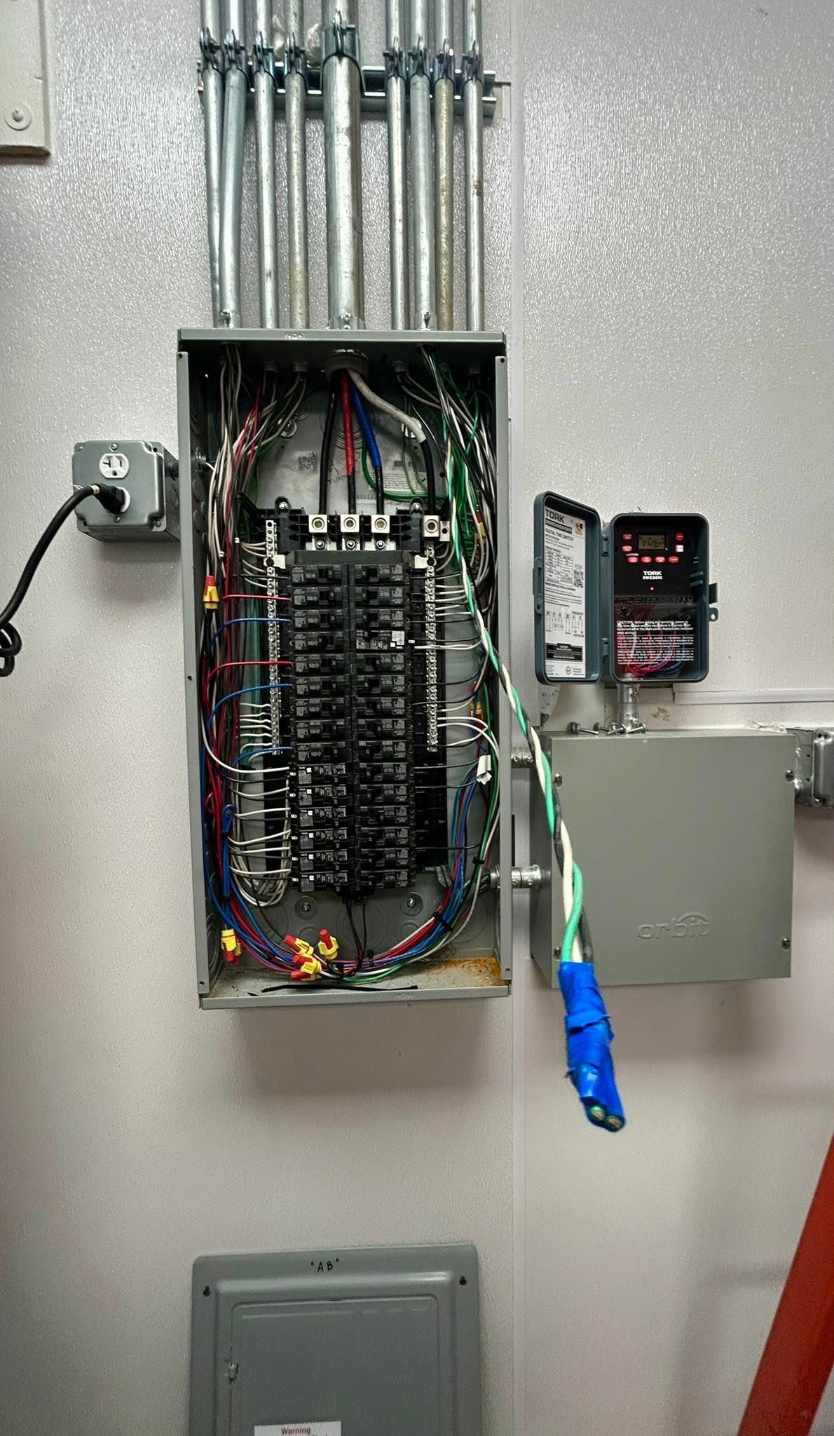 An open electrical panel mounted on a wall with conduit piping, a small digital display, and exposed wiring.