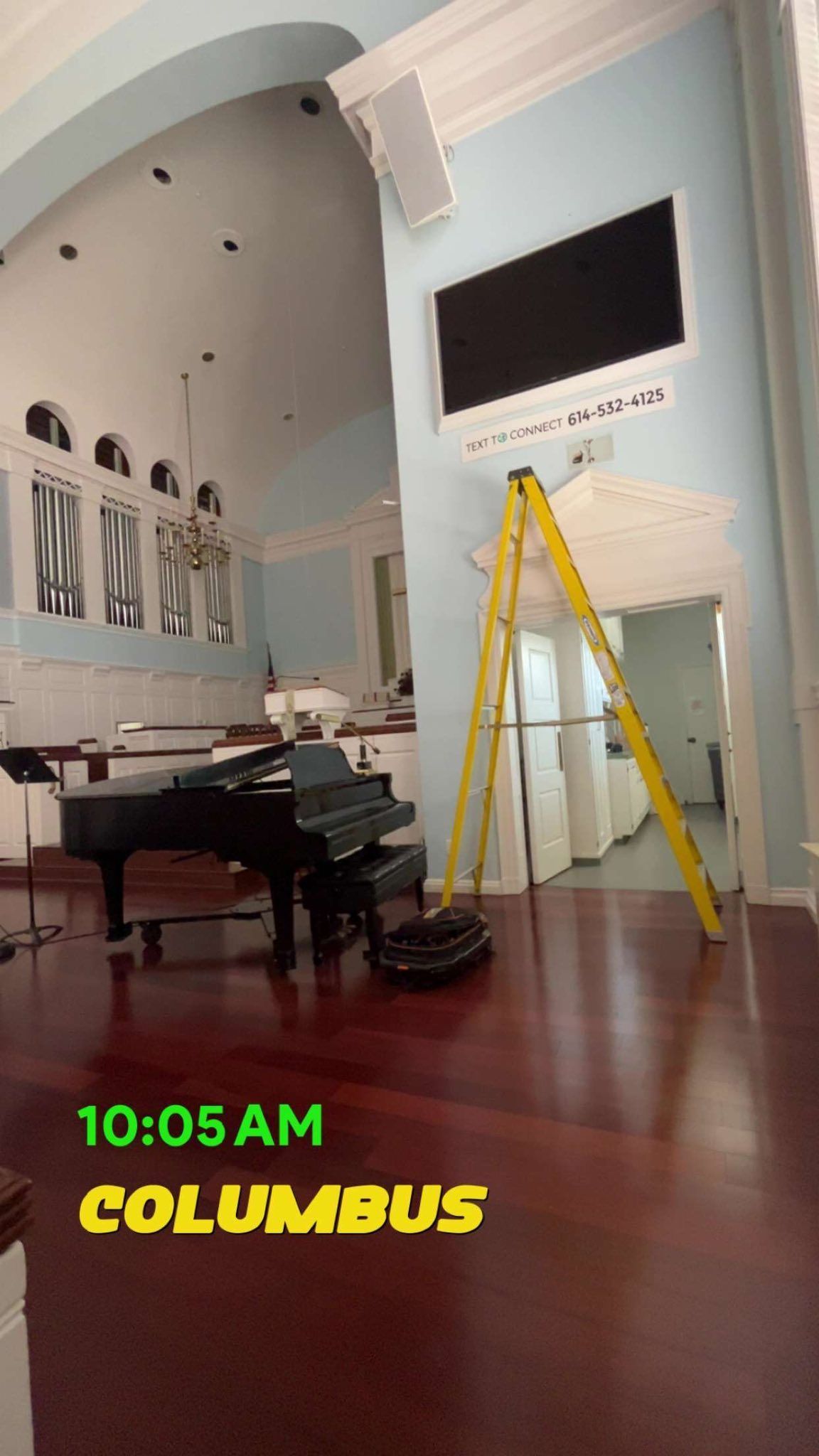 A bright interior with a black grand piano, a tall yellow step ladder, and blue walls. Text overlays: 10:05 AM, COLUMBUS.