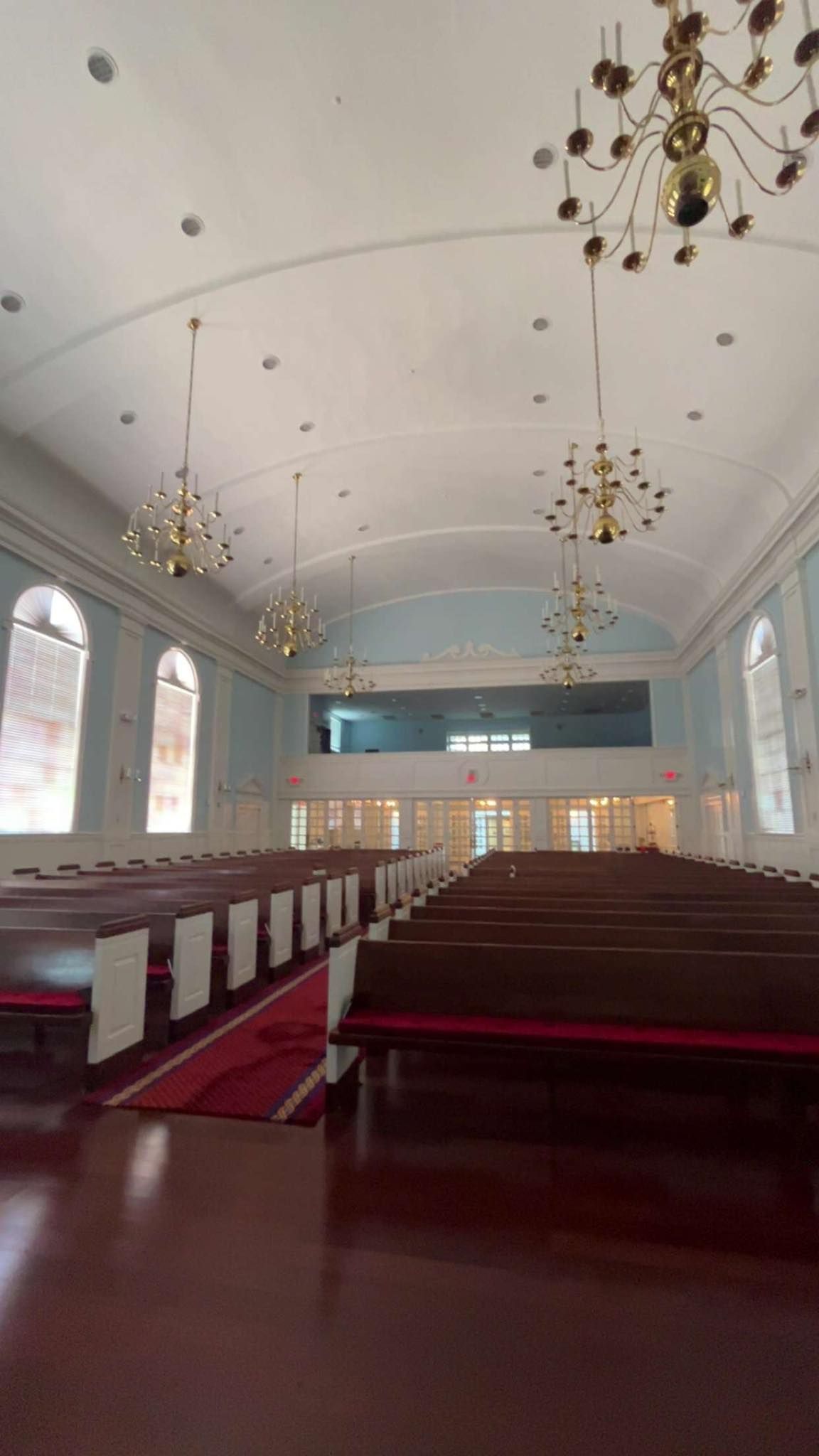 Interior of a church with rows of wooden pews, a red carpet runner, arched windows, and brass chandeliers.