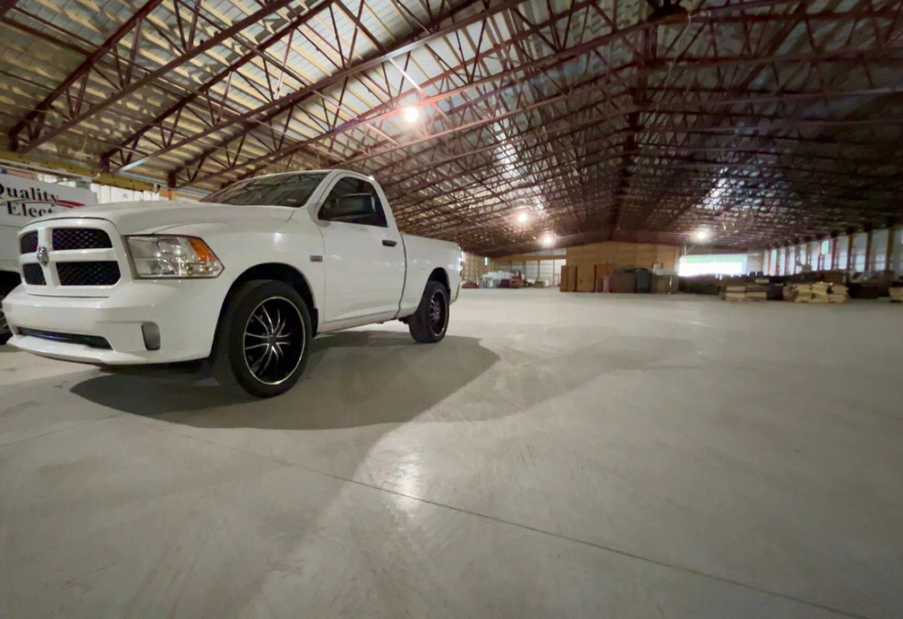 A white pickup truck parked on a large concrete floor inside a wide, open-truss industrial warehouse.