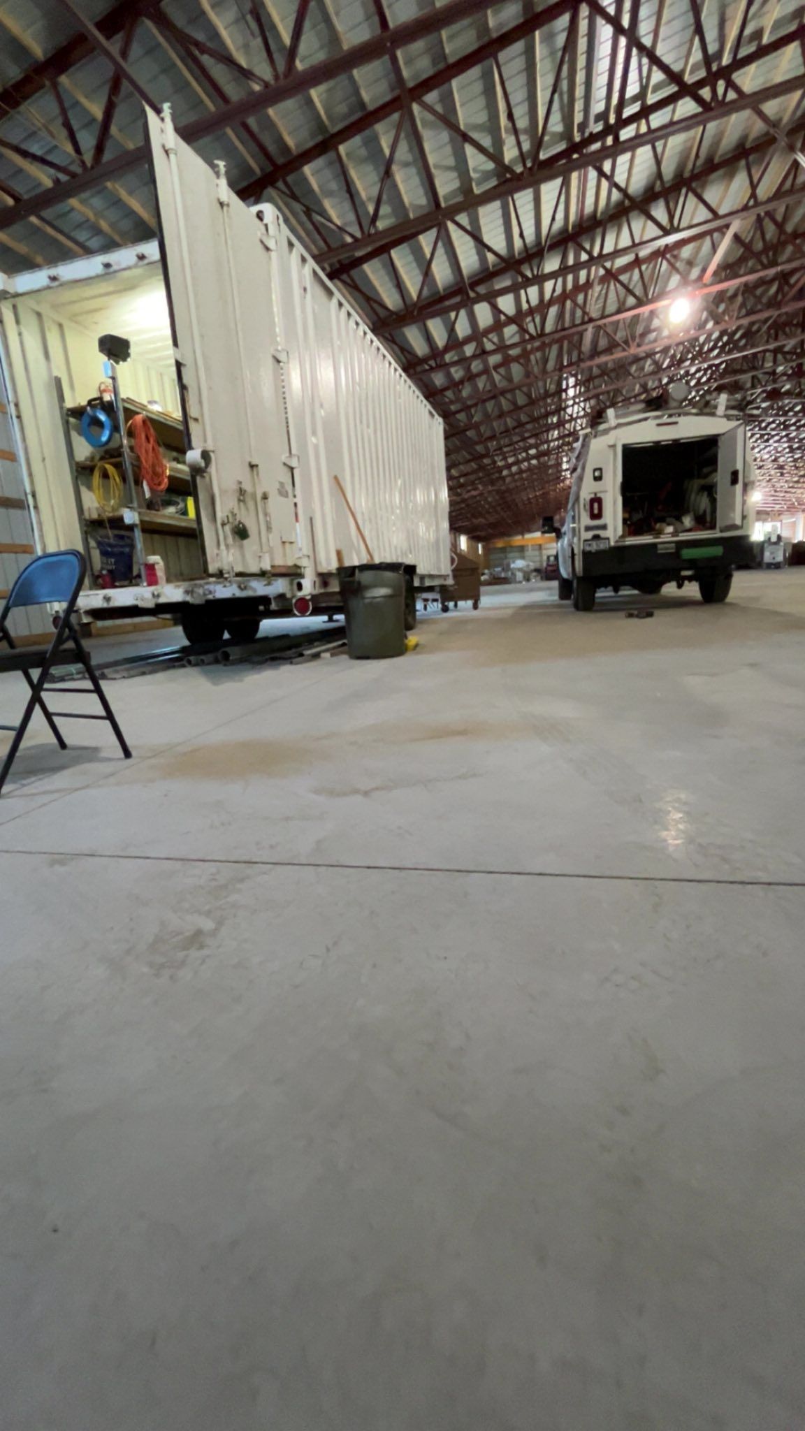 A white trailer and a service truck parked inside a large, open warehouse with a high metal-truss ceiling.