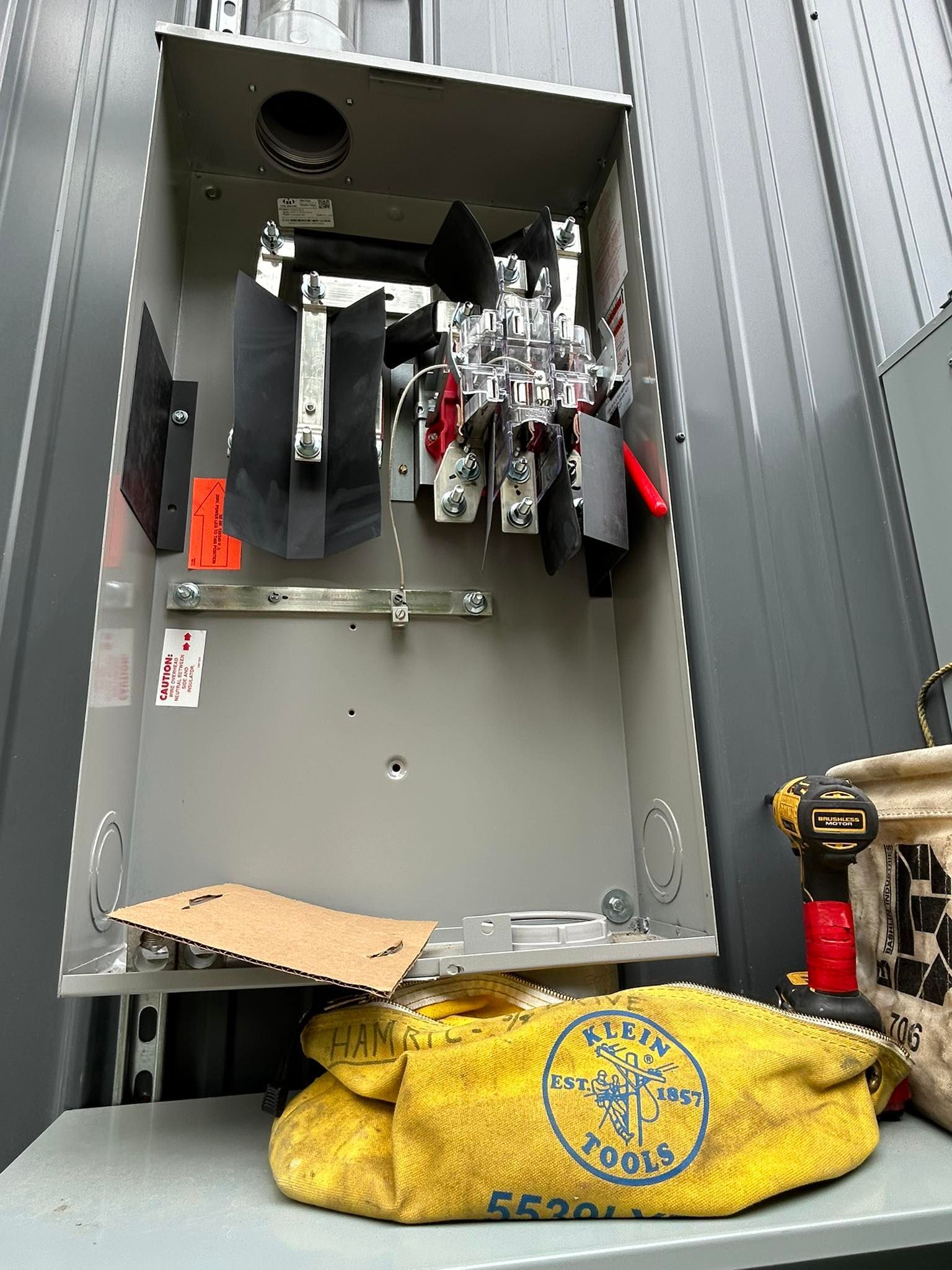 An open, gray electrical meter socket mounted on a corrugated metal wall with a tool bag and drill resting below.
