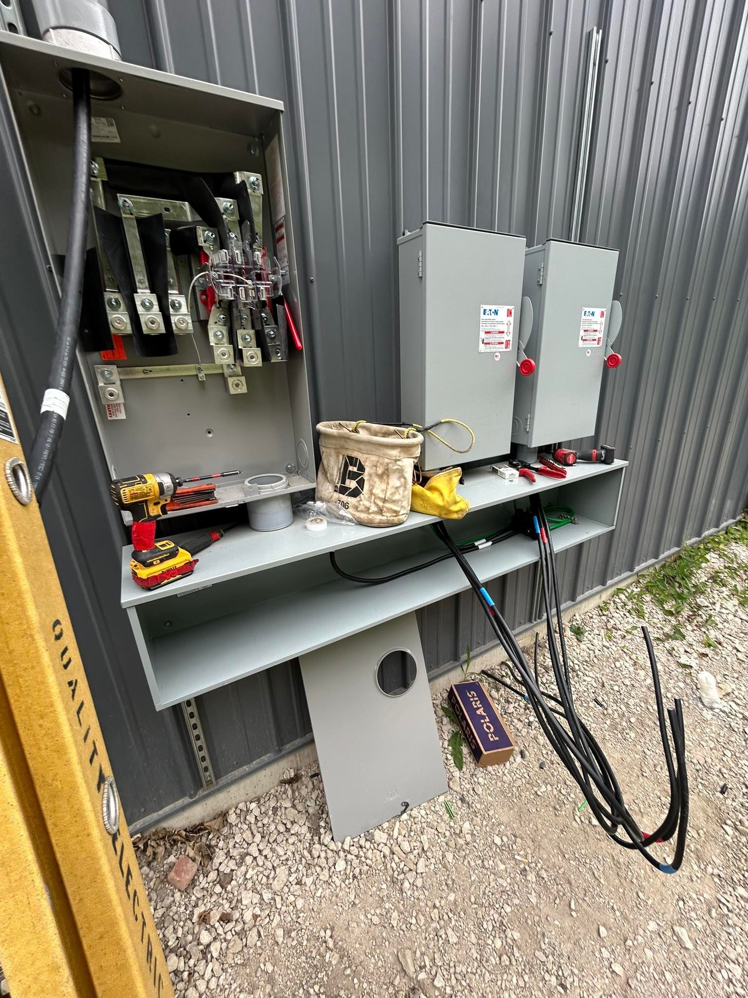 An open electrical panel and two mounted disconnect switches on a corrugated metal wall, with tools and wires on a shelf.