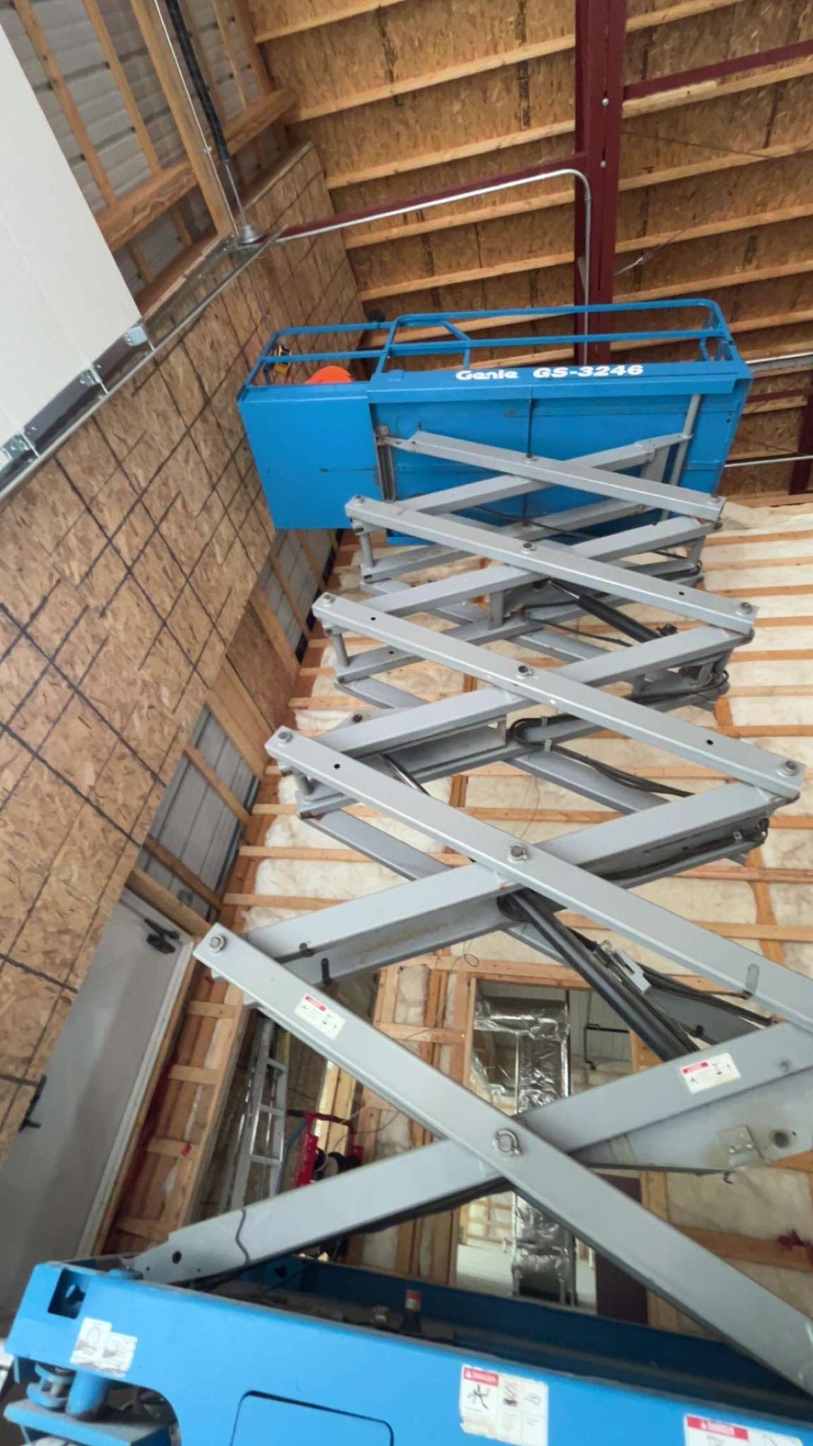 A blue Genie scissor lift fully extended vertically inside a structure with wooden walls and ceiling trusses.