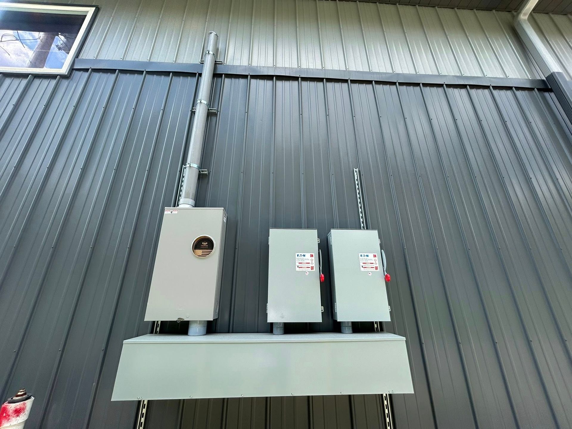 An electrical meter box and two disconnect switches mounted on a dark, vertical ribbed metal wall.