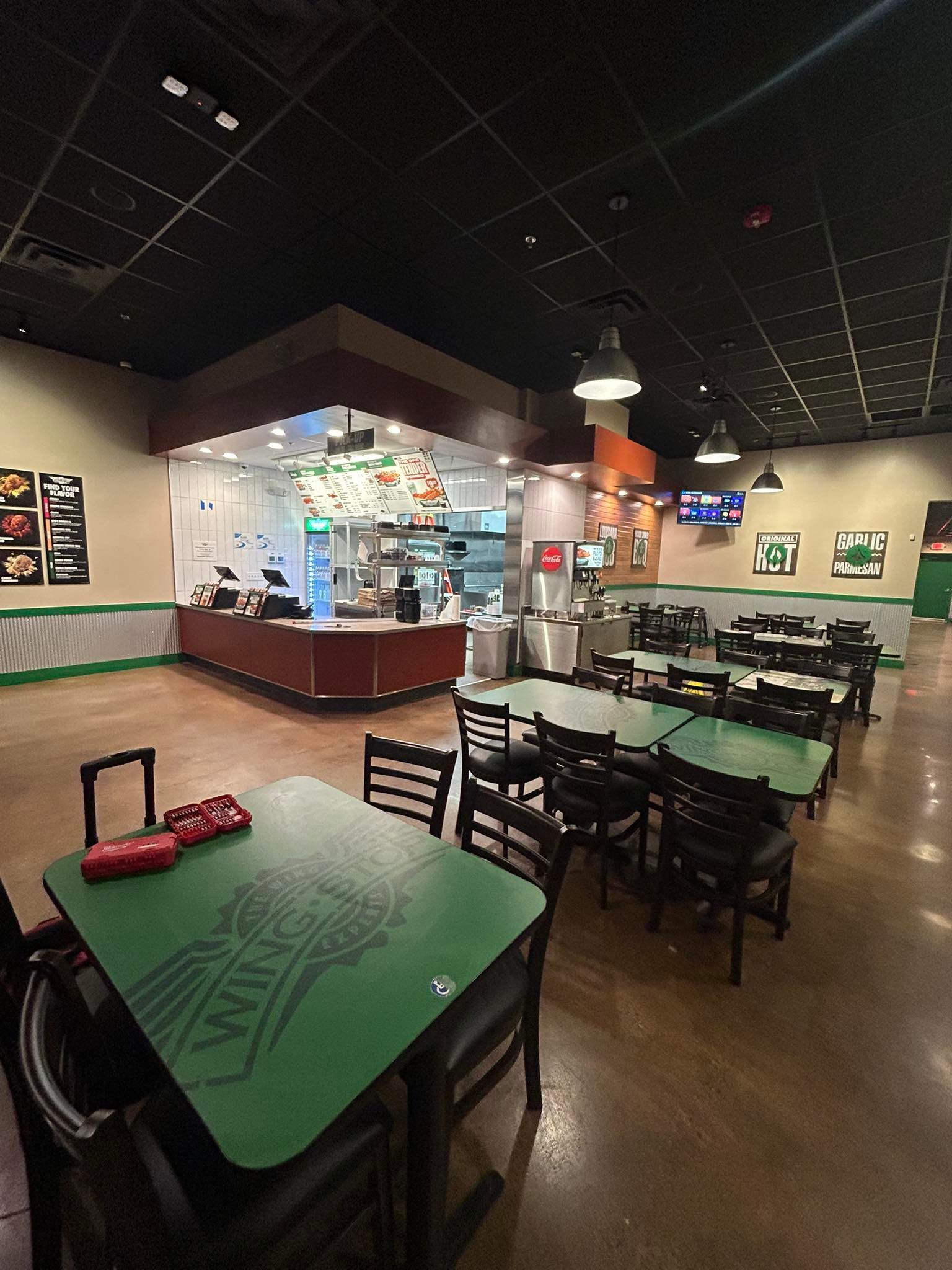 A dining area with green tables and black chairs, featuring a service counter in the background under warm lighting.