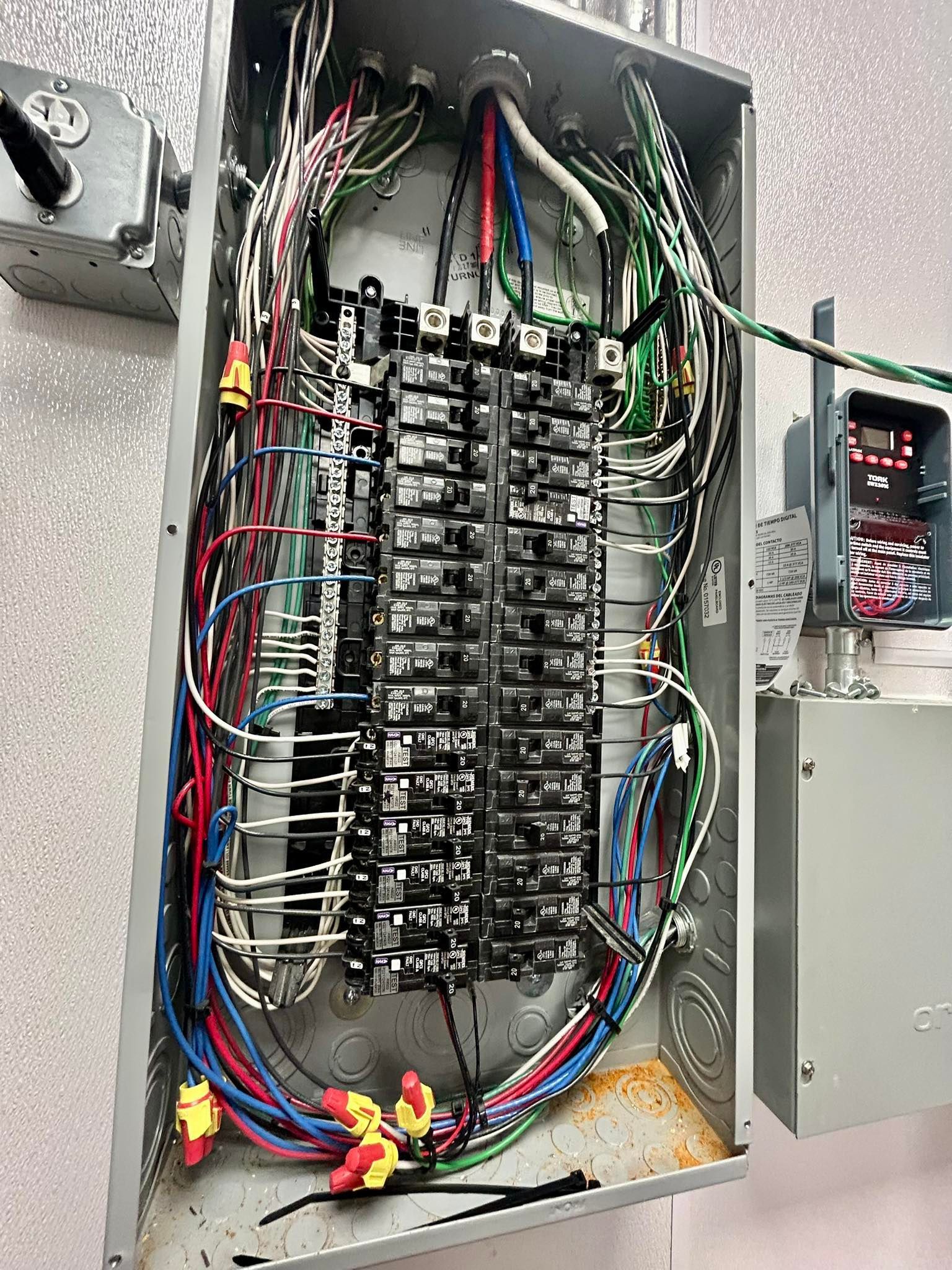 An open electrical panel with multicolored wiring connected to various circuit breakers mounted on a wall.