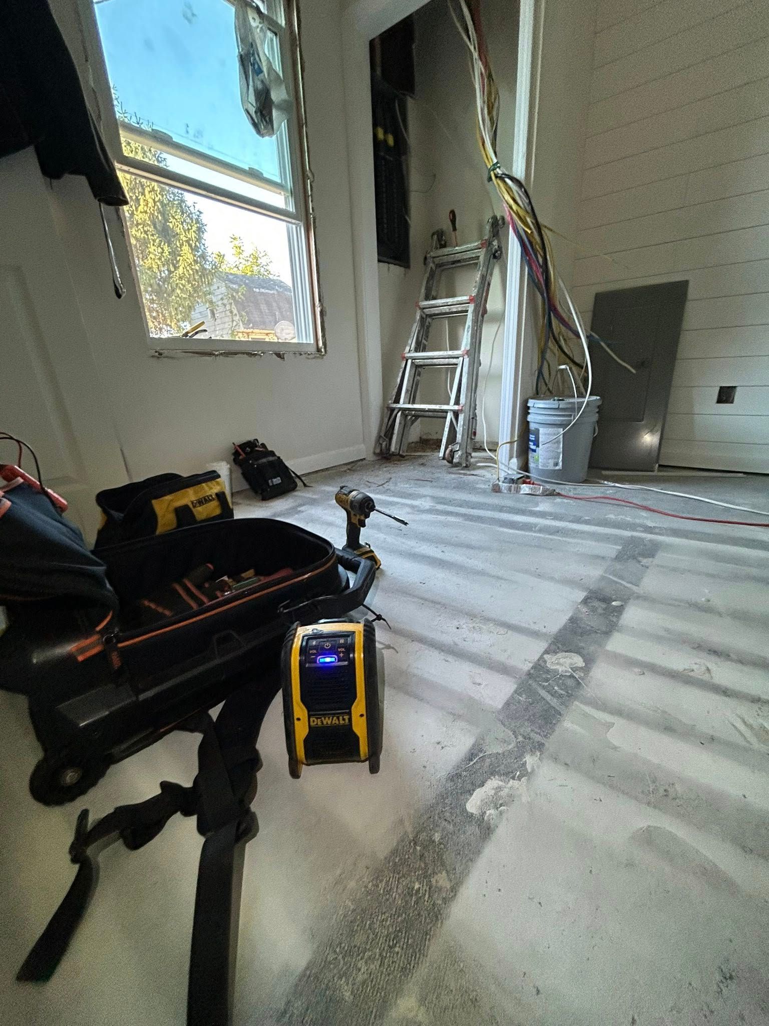 Construction tools, a work light, and a ladder inside an unfinished room with exposed wiring and a window.