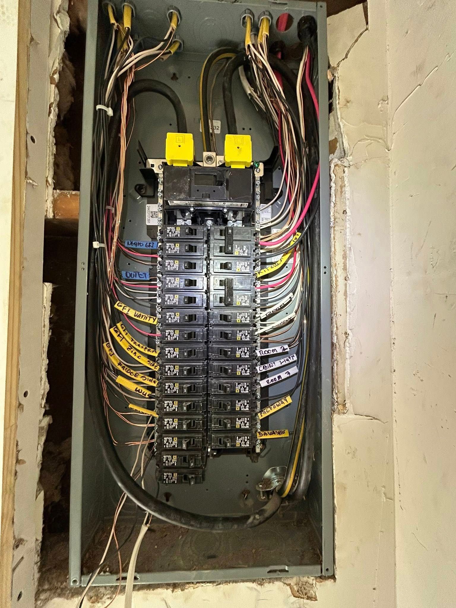 An electrical panel with numerous breakers, wires, and yellow plastic connectors, mounted inside a wall opening.