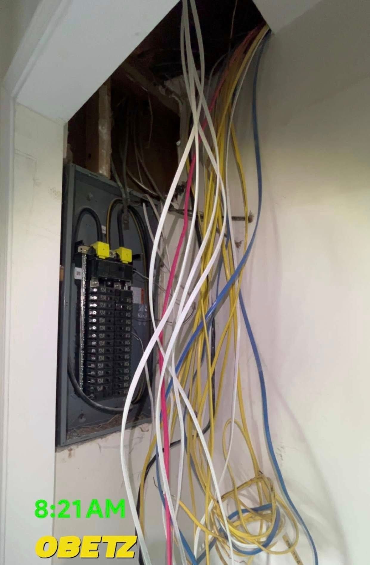 Electrical panel box with exposed, messy wiring hanging from an open wall cavity above it.