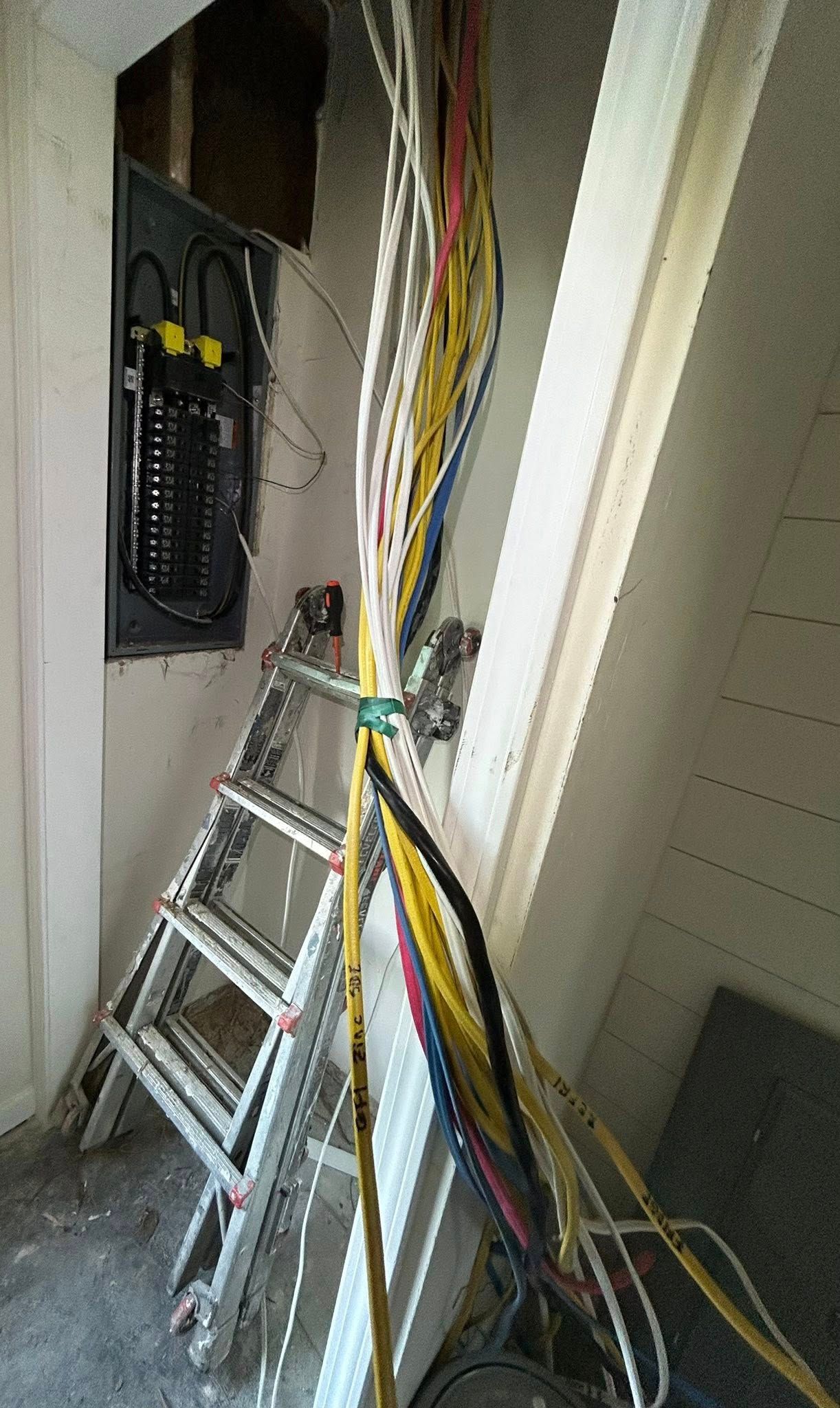 An open electrical panel on a wall near a stepladder with a large bundle of multicolored wires hanging in the foreground.