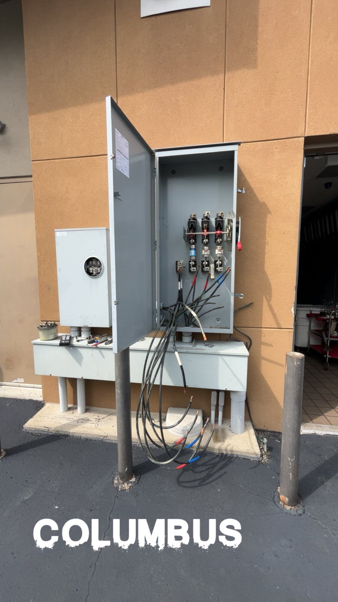 An open electrical utility cabinet mounted on an exterior wall with exposed wiring and protective metal bollards.