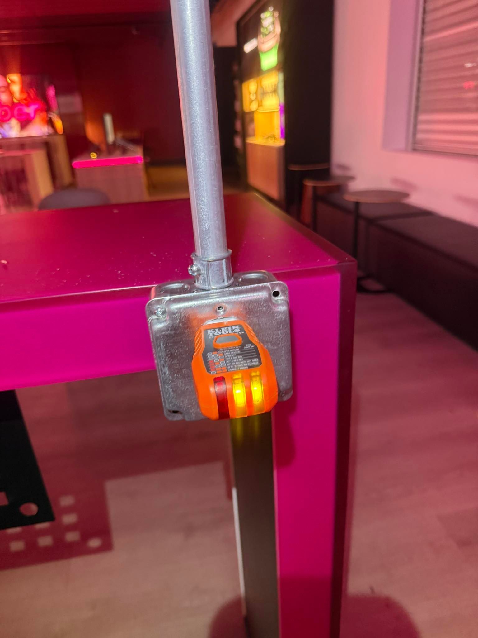 An orange electrical tester plugged into a metal utility box mounted to a pink table leg in a dimly lit room.