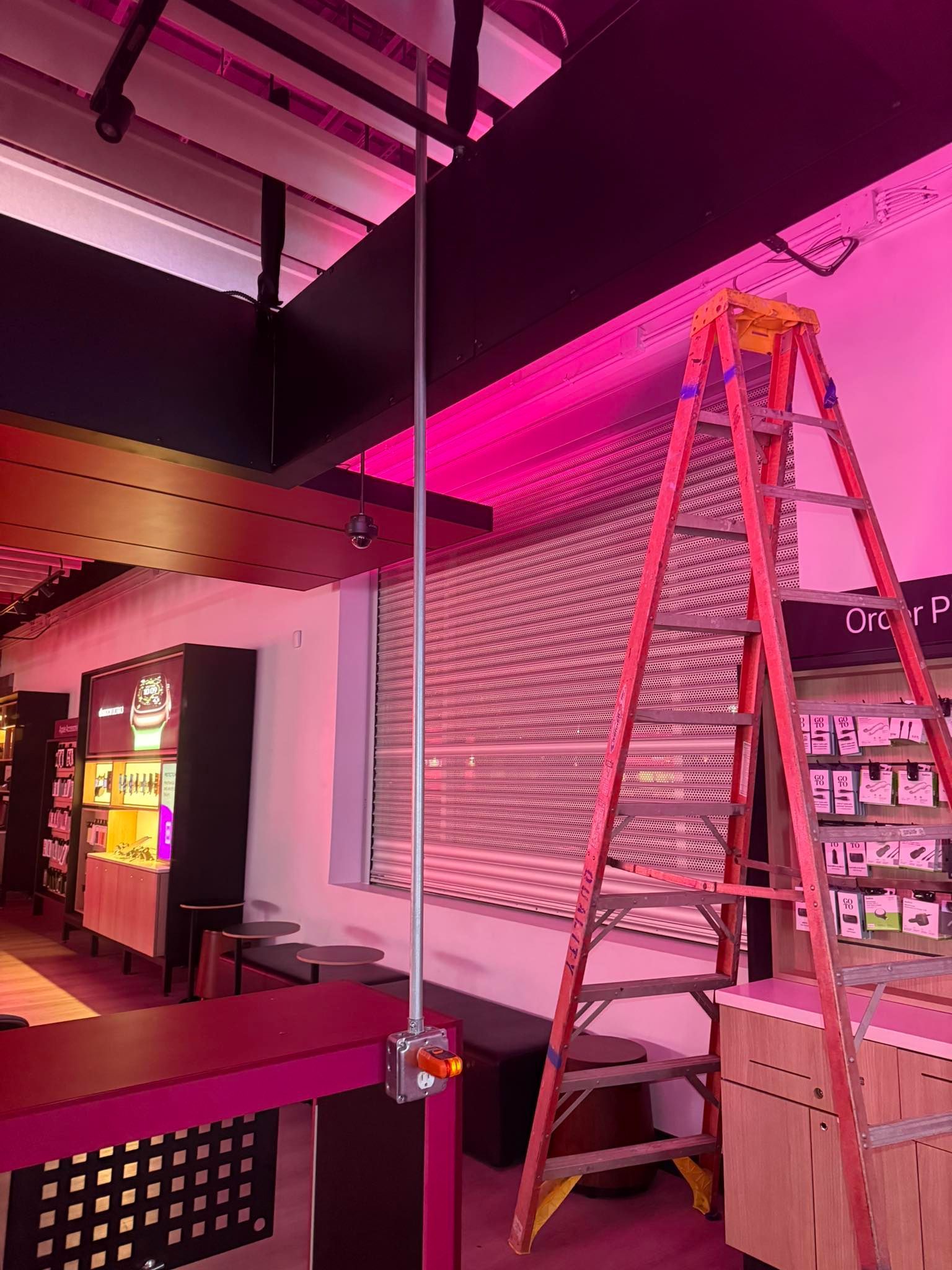 A tall, red A-frame ladder stands in a brightly lit room with pink lighting, next to a metal conduit on the wall.