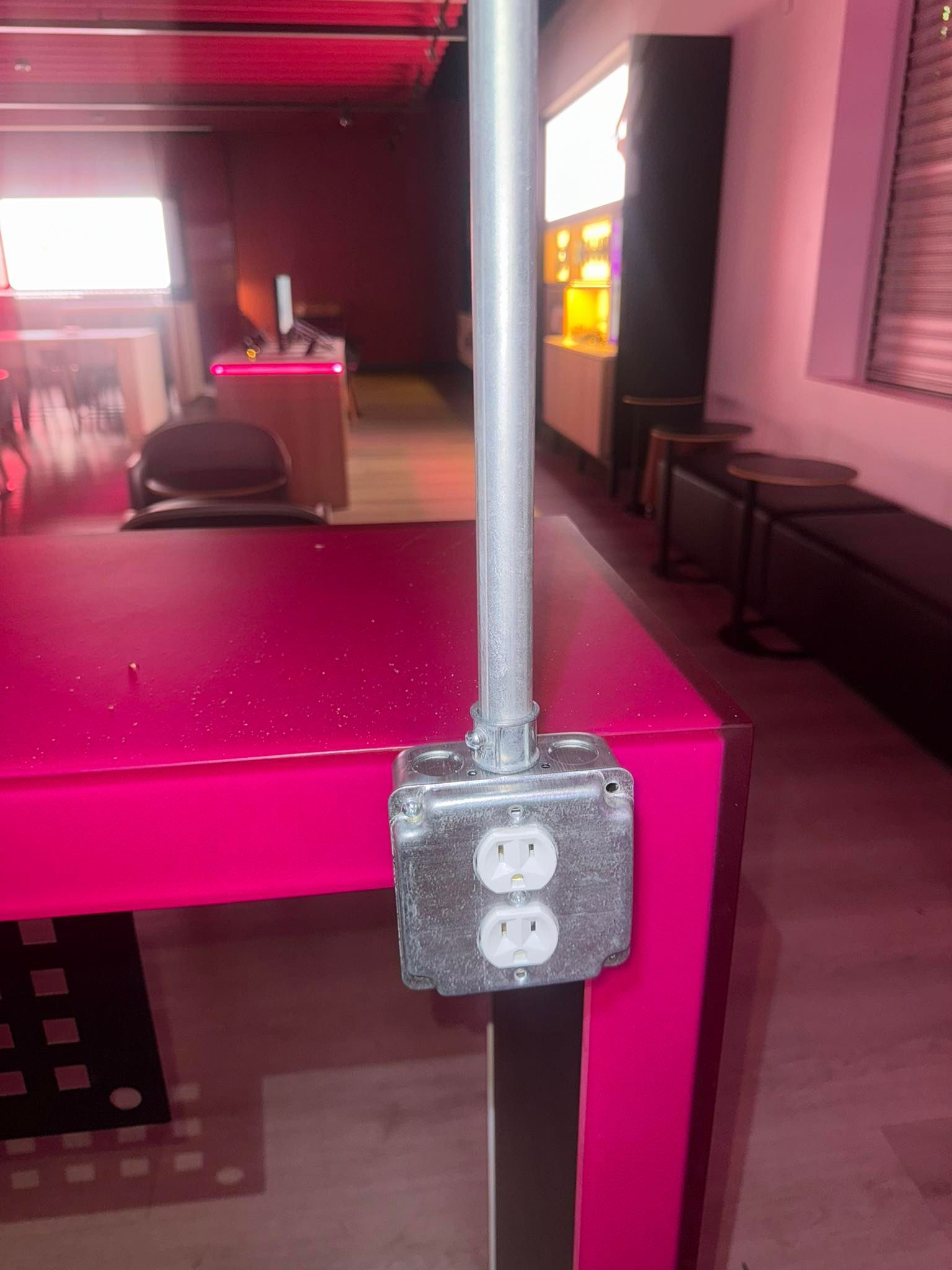 A metal electrical outlet box mounted on the edge of a vibrant pink table in a room with dim red lighting.