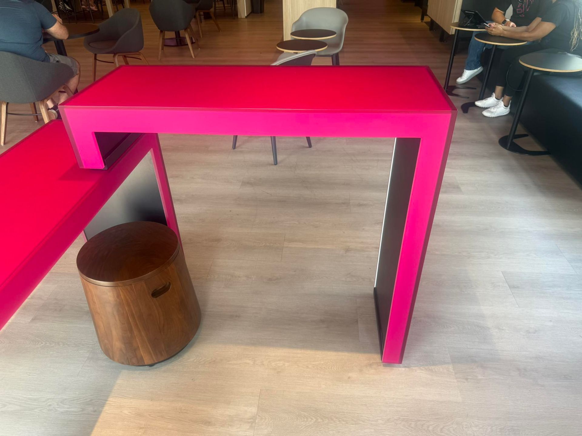 A bright pink, L-shaped modern standing desk with a wooden stool underneath in a contemporary cafe setting.