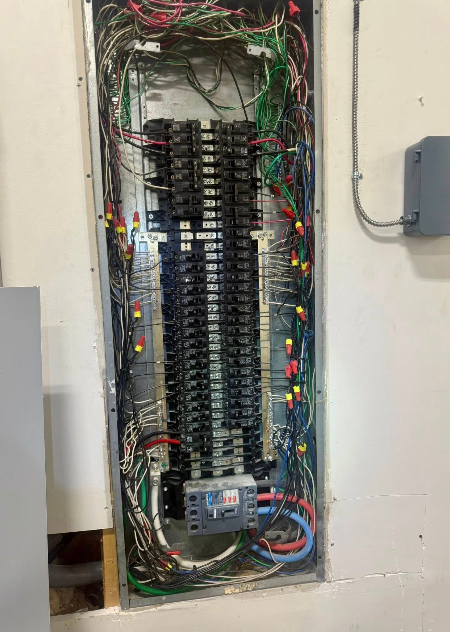 A large, vertical electrical breaker panel mounted on a wall, showing numerous circuit breakers and colored wiring.