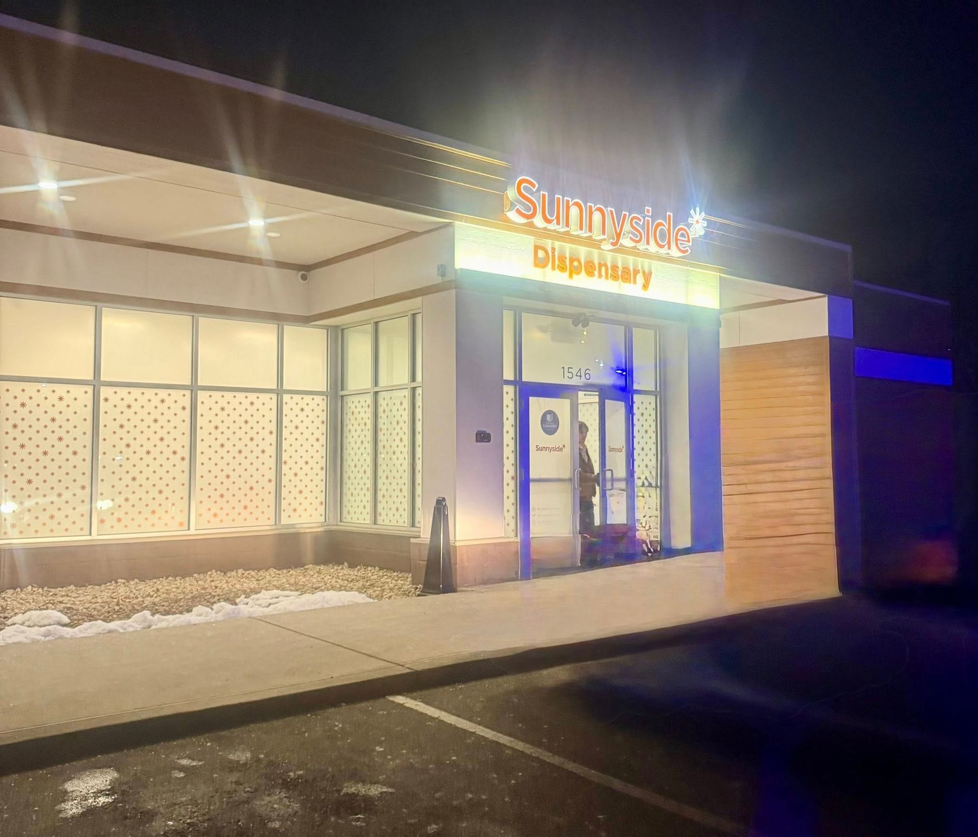 Exterior of a Sunnyside Dispensary storefront at night, featuring a glass entrance, wood paneling, and interior lighting.