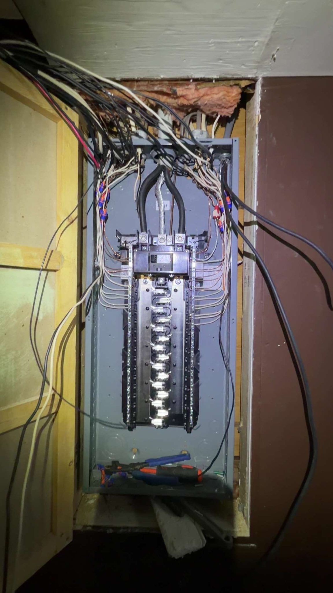 An open residential electrical service panel showing wire connections to the bus bars, with tools resting at the bottom.