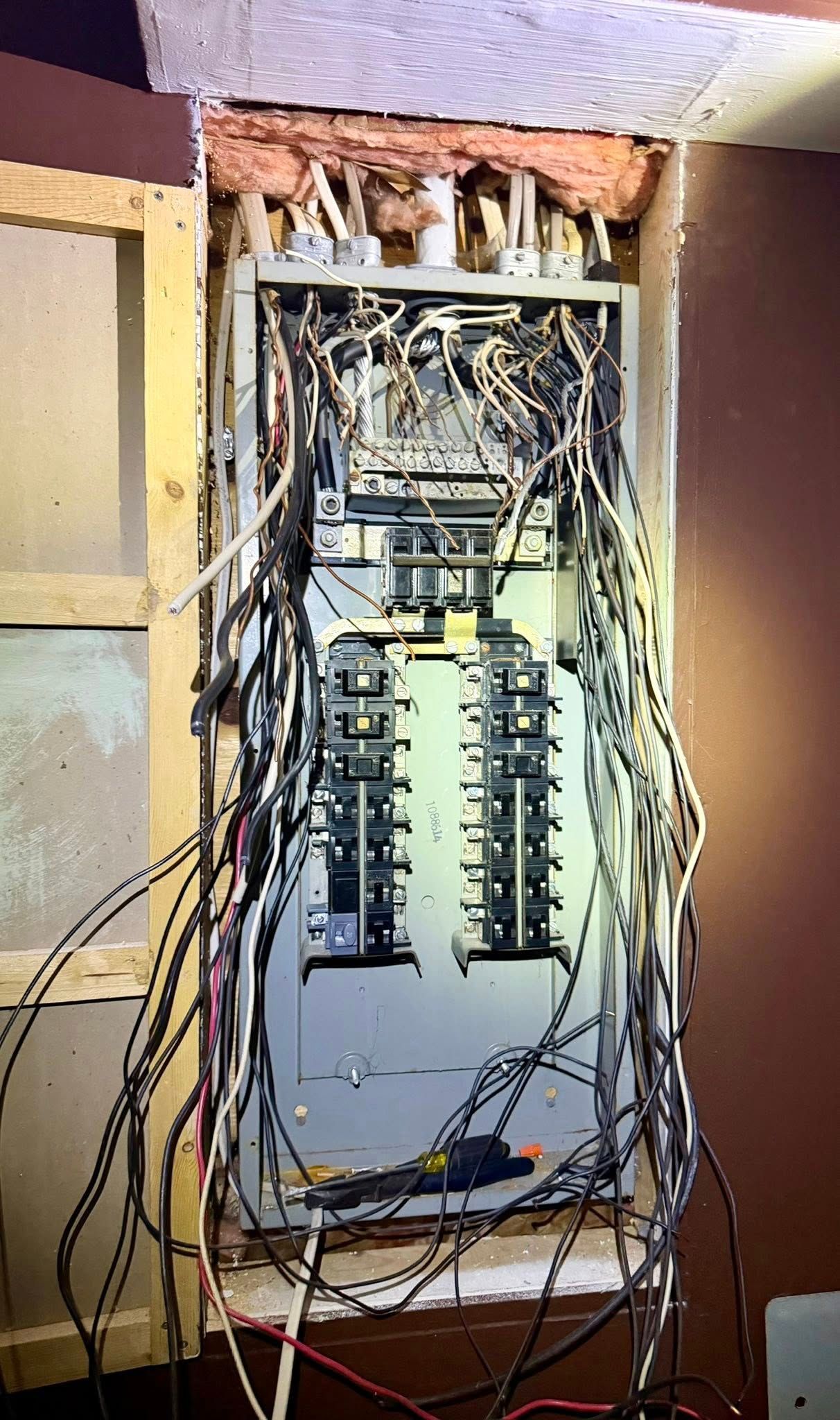 An open electrical panel box mounted in a wall cavity, with numerous wires connected to breakers and bus bars inside.