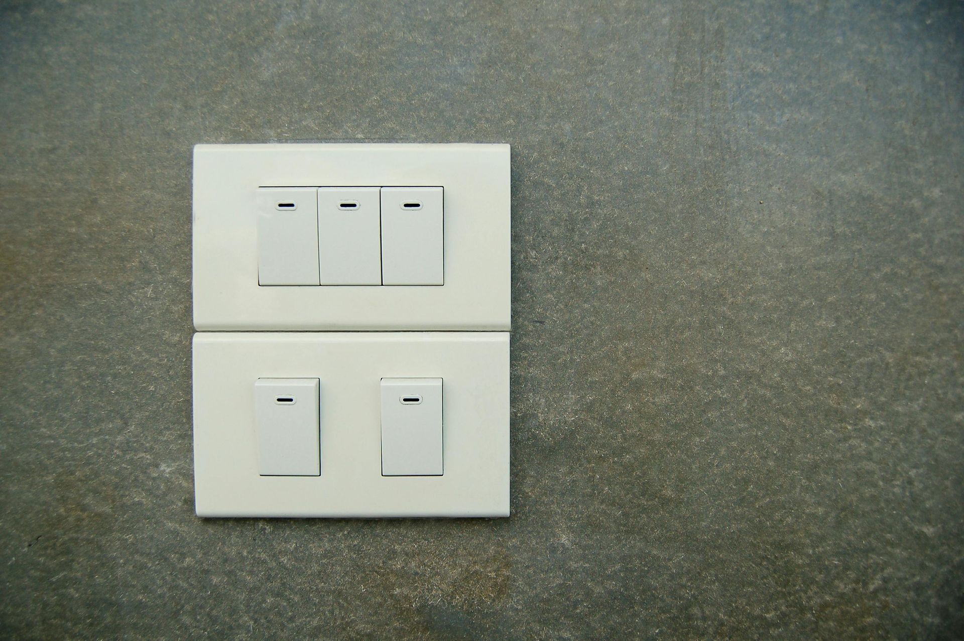 A set of five light switches in two white rectangular panels mounted on a dark, textured gray wall.