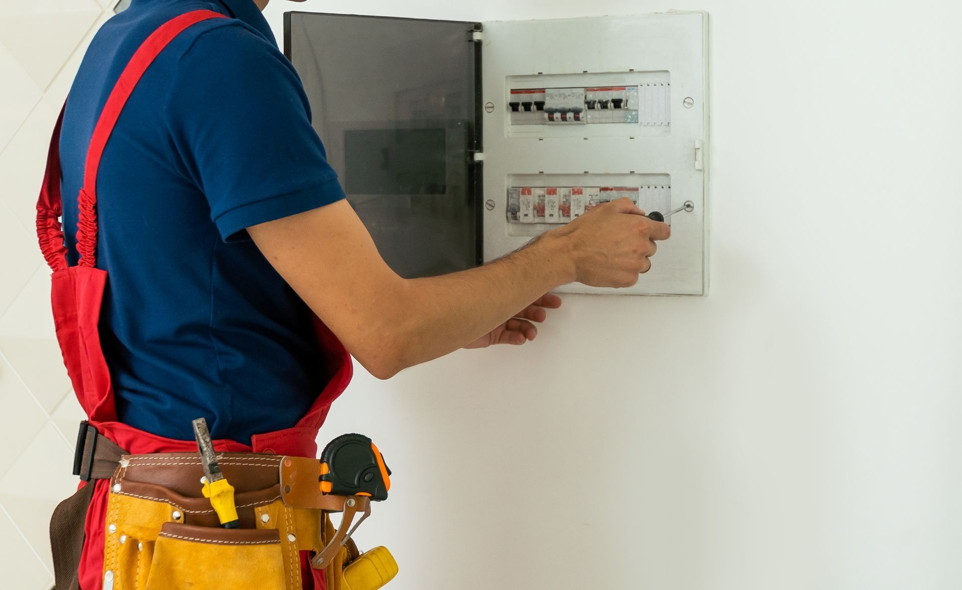 An electrician in a blue shirt and red overalls uses a screwdriver to work on an open electrical service panel.